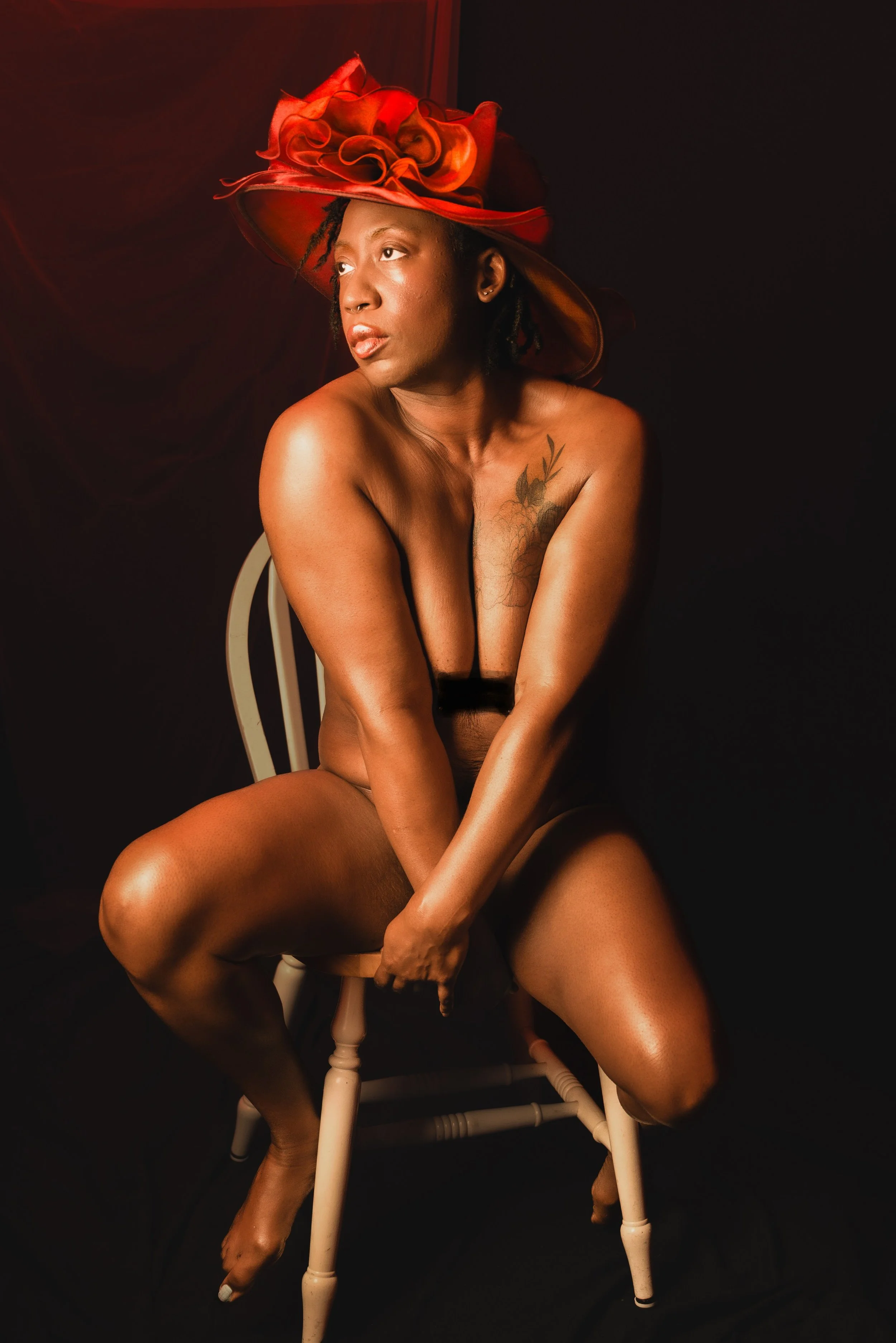 A woman sitting nude on a white chair, wearing a large red hat with decorative red ribbons, posed against a dark background.