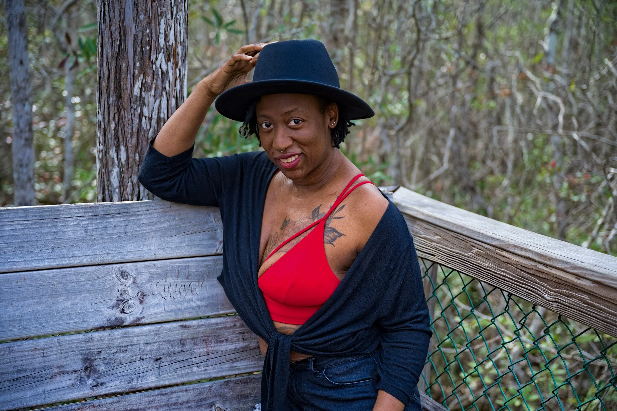 A woman wearing a black hat, a red tank top, and a black cardigan stands outdoors next to a wooden fence and trees, smiling and touching her hat.