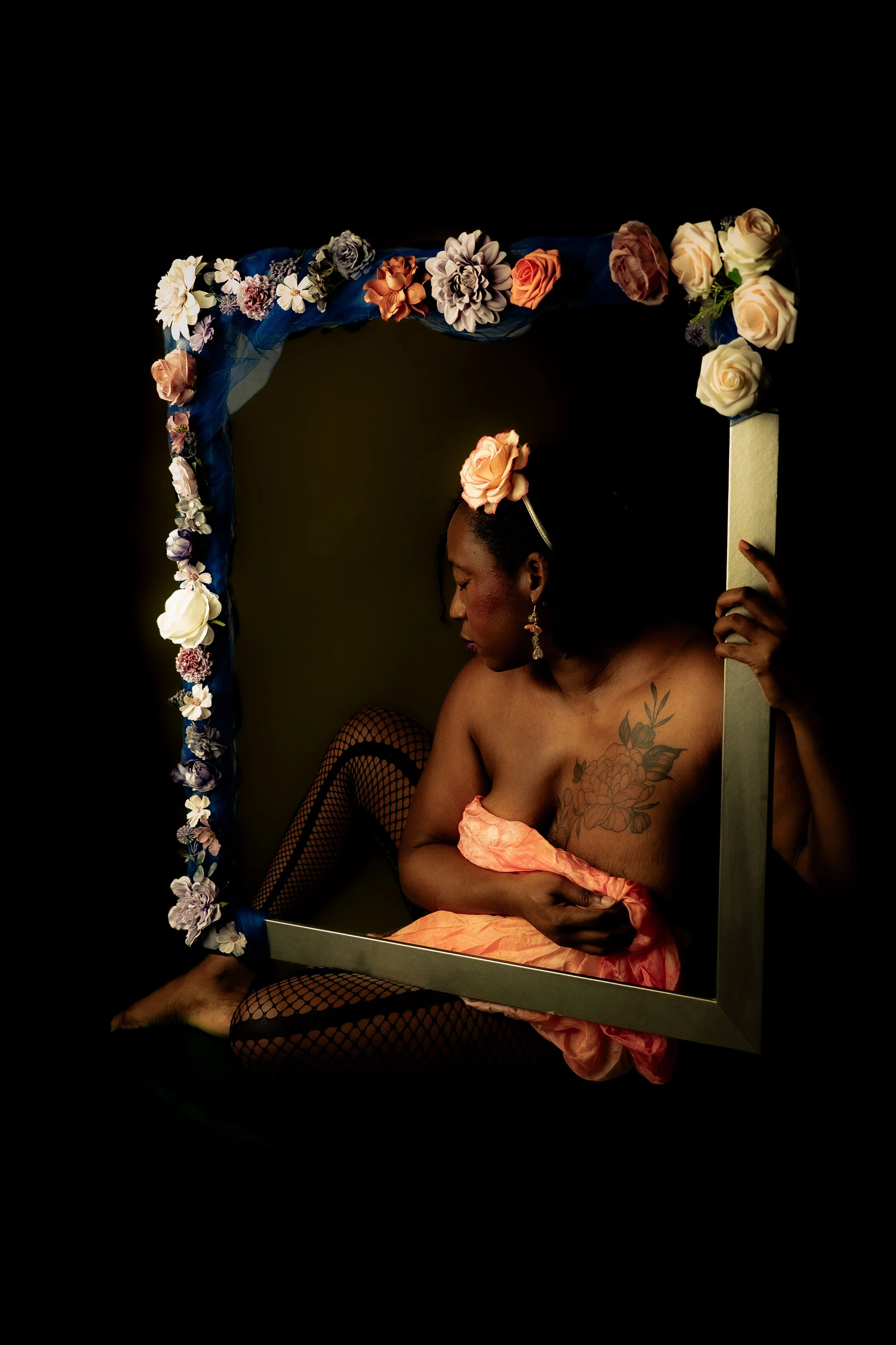 A woman with a flower on her head sitting inside a photo frame decorated with flowers, wearing fishnet stockings and a pink garment, looking down