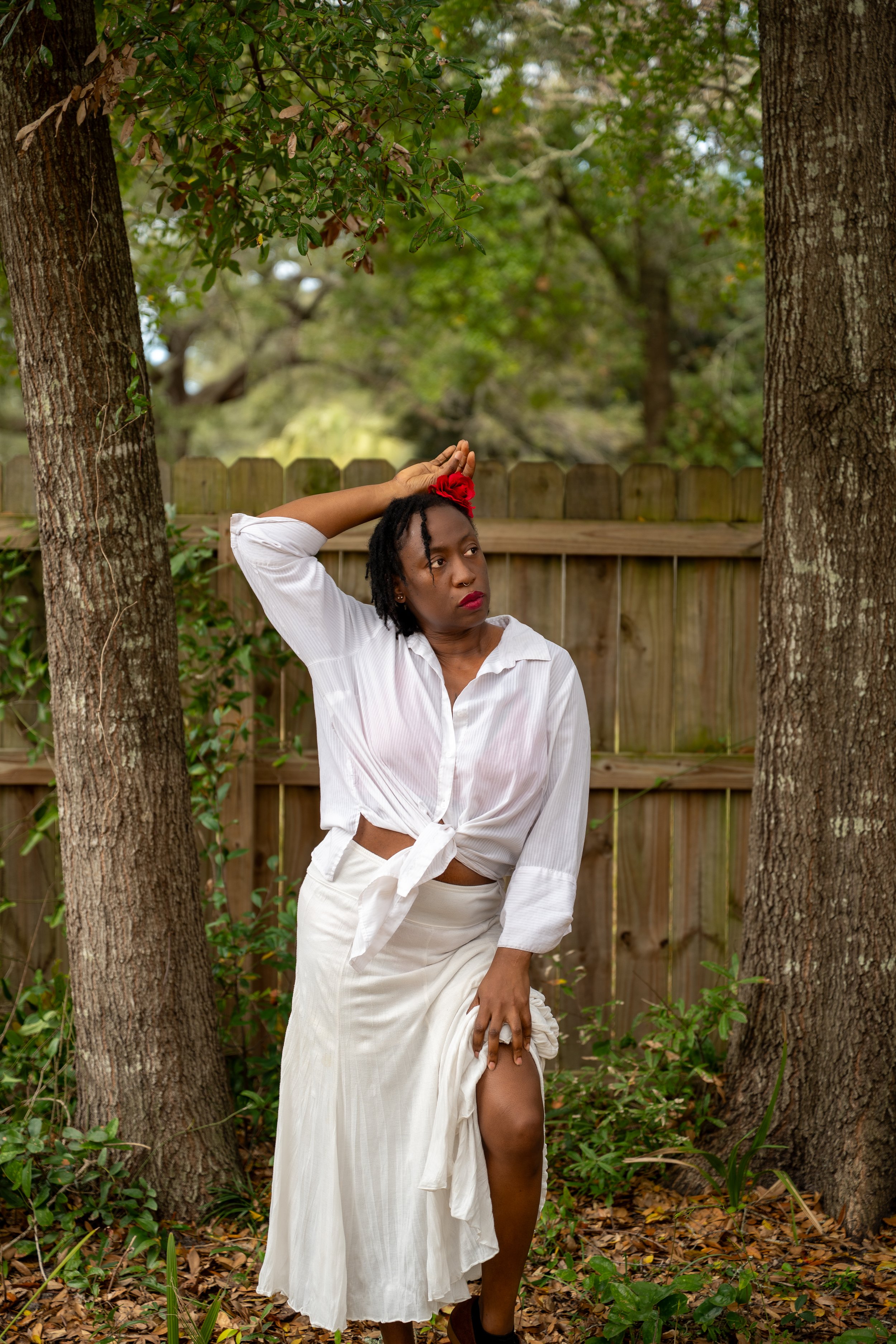A woman standing outdoors in a wooded area with a wooden fence behind her. She is wearing a white tied-up shirt, a long white skirt, and a red flower in her hair. She is holding up her skirt with one hand and resting her other hand on her head, looki