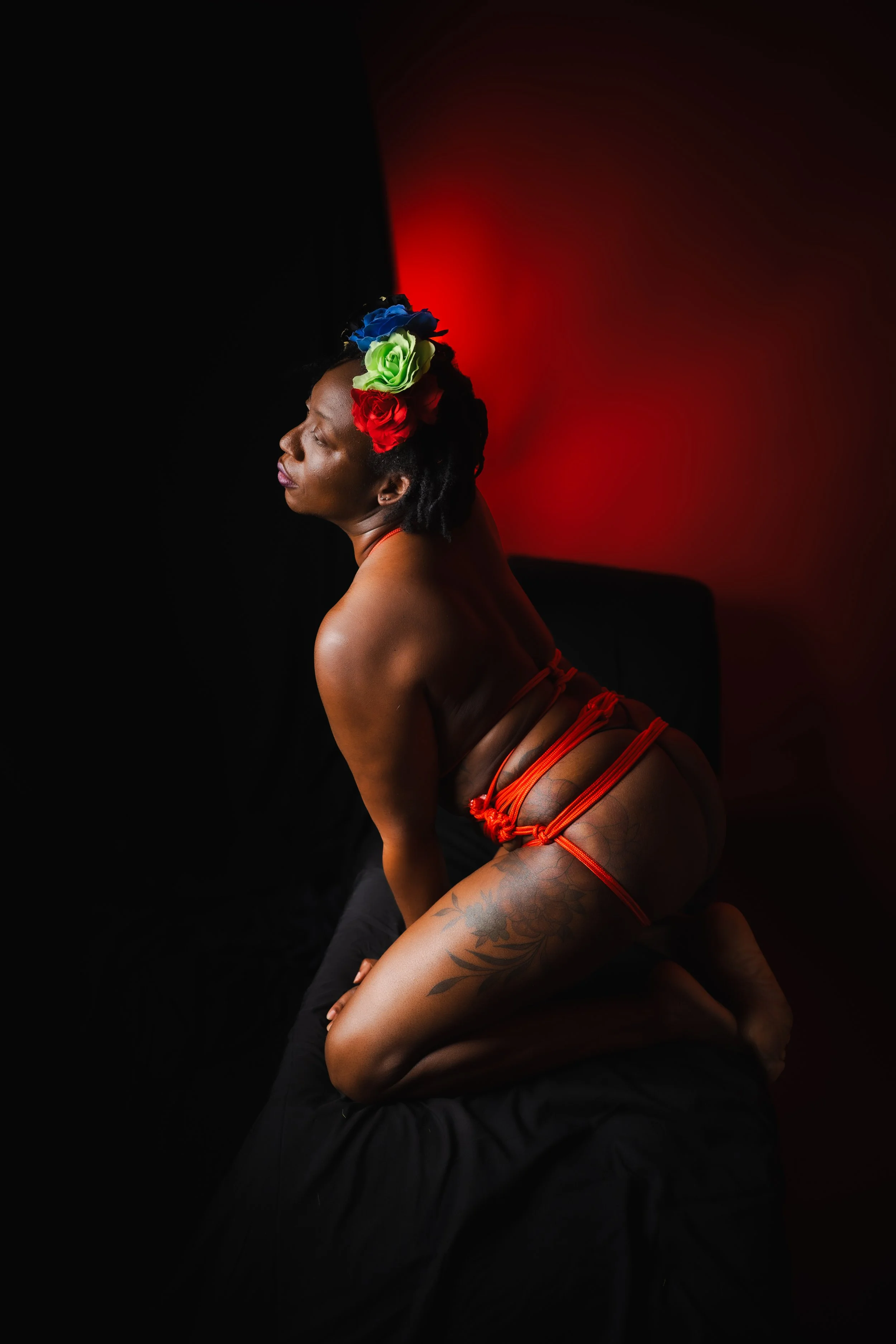 Shibari portrait of a tattooed woman in red rope harness with a vibrant multicolor floral crown, kneeling in profile against a split black and red background.