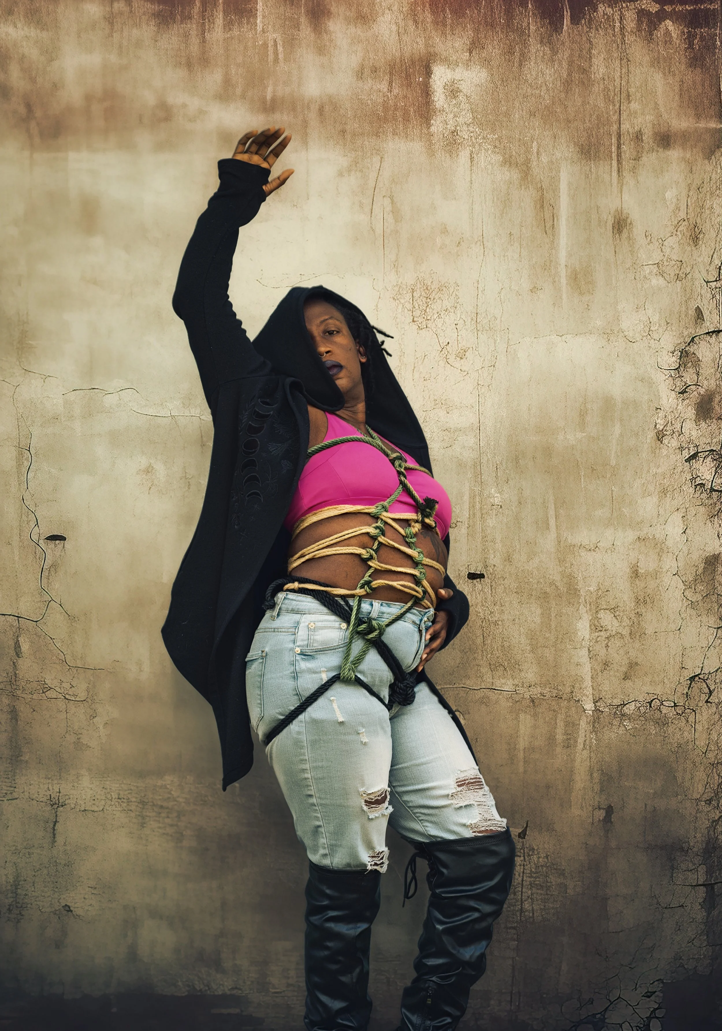 A woman with dark skin and dreadlocks poses against a cracked, weathered wall. She is wearing a pink crop top, distressed jeans, a black jacket with hood, and over-the-knee black boots. Her hands are tied with ropes, and her left hand is raised above her head.