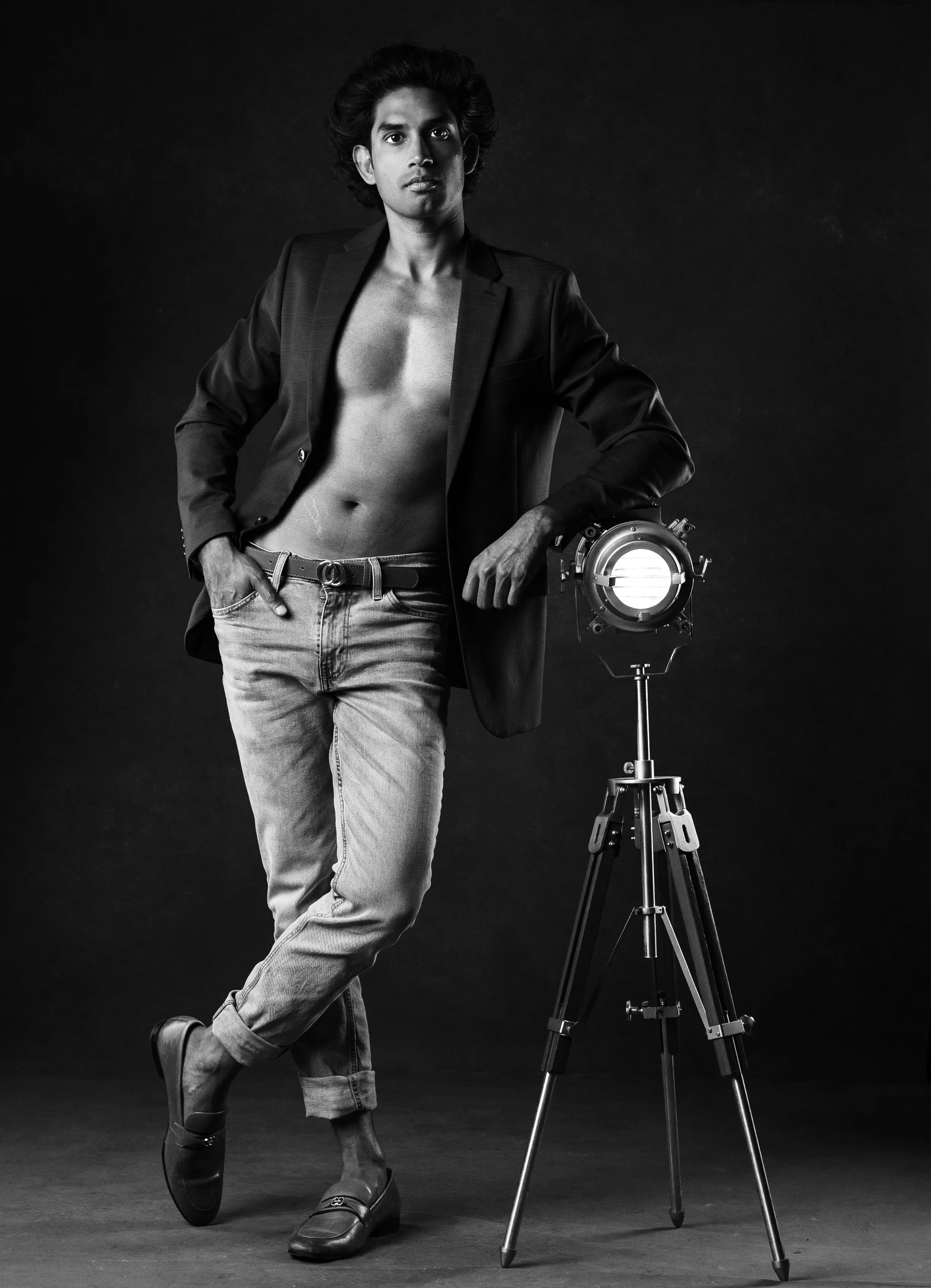 Black and white photo of a young man with dark, curly hair, shirtless and wearing a suit jacket and jeans, standing with one hand on his hip next to a studio light on a tripod against a dark background.  Boudoir for Men.  Male Boudoir. Gentlemen Boud