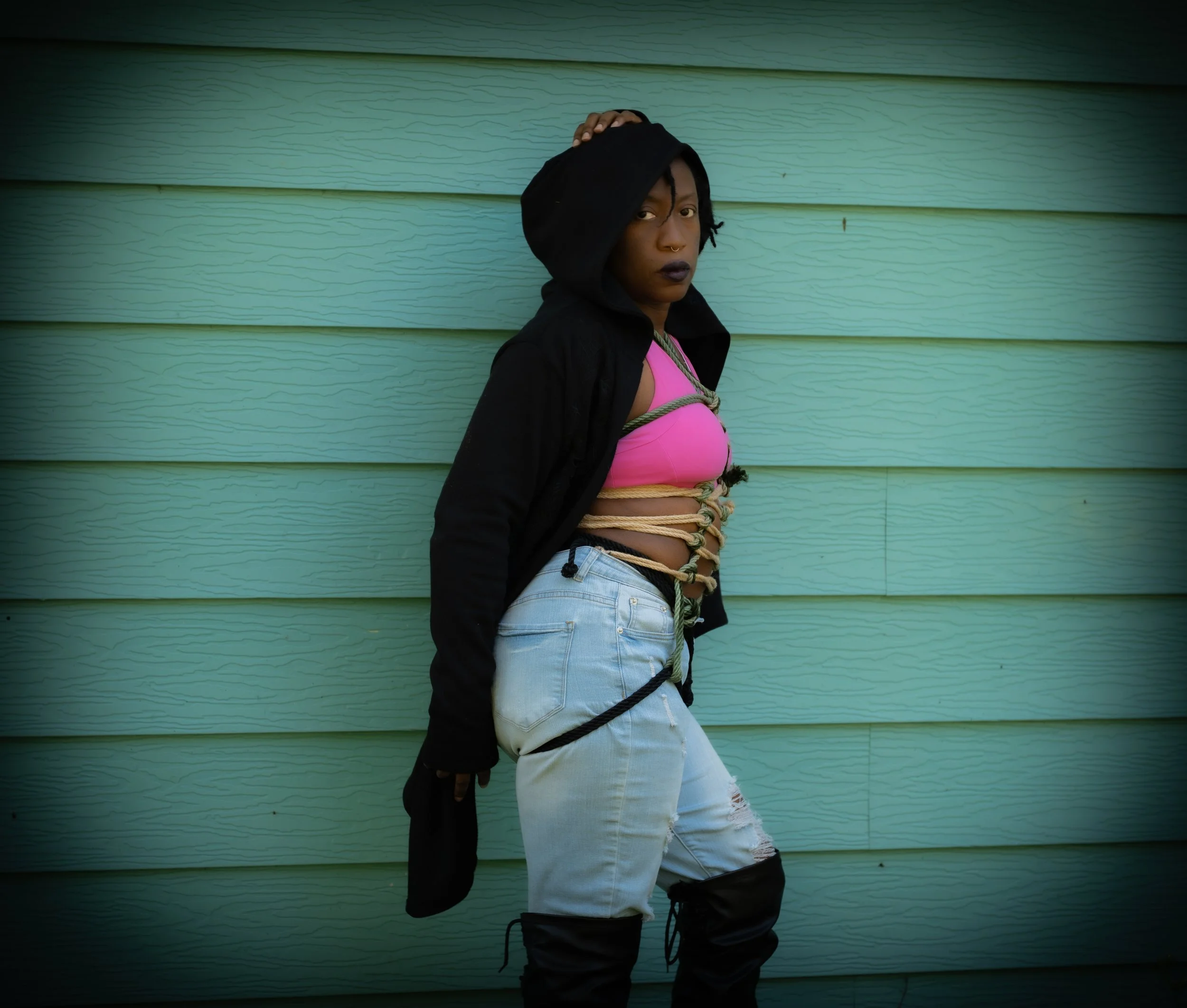 Young woman with dark lipstick and nose ring leaning against a light green wooden wall, wearing a black hooded jacket, pink top, ripped jeans, and black boots with her hand on her hood.