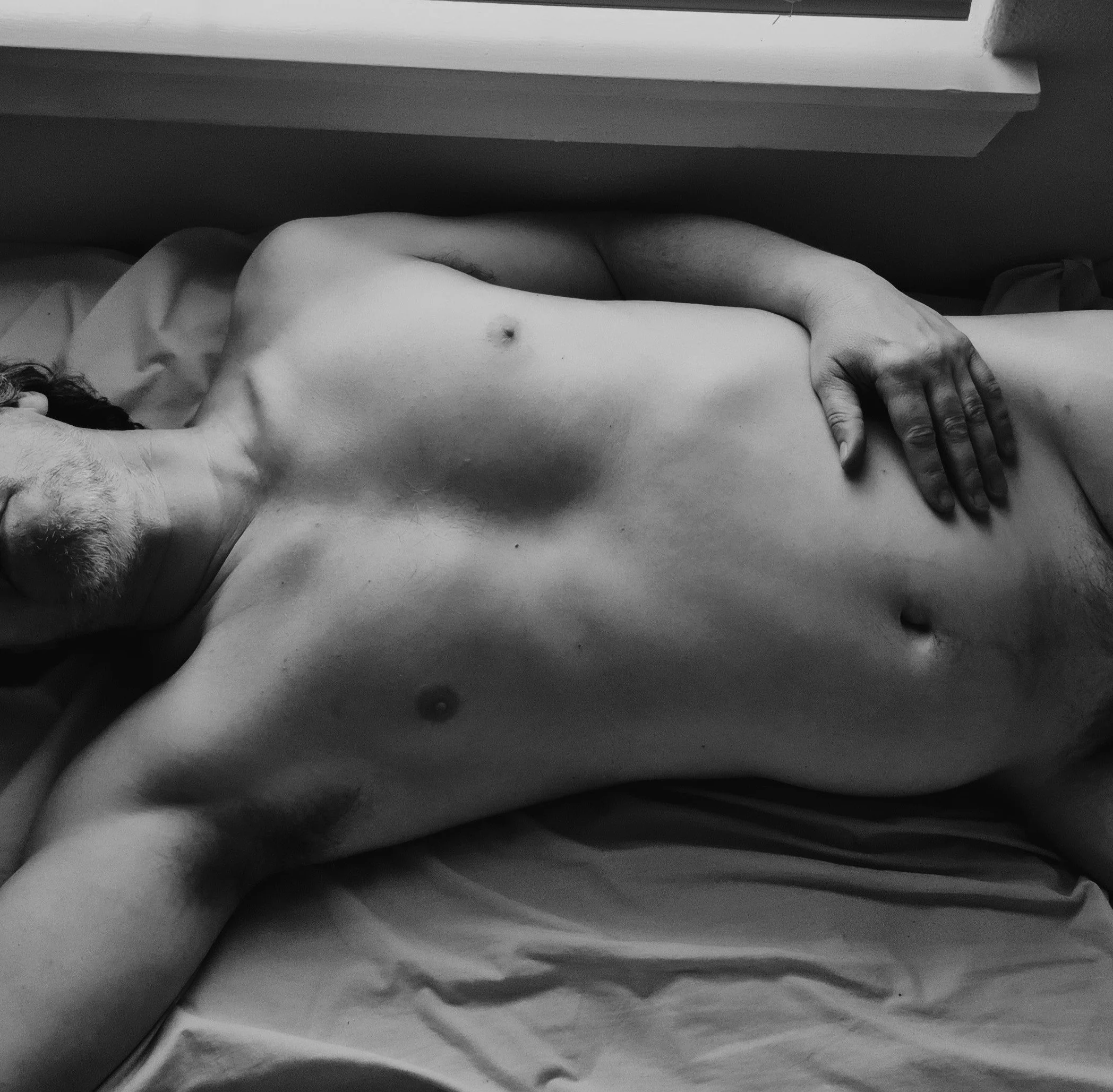 A shirtless man lying on a bed with his hand resting on his stomach.