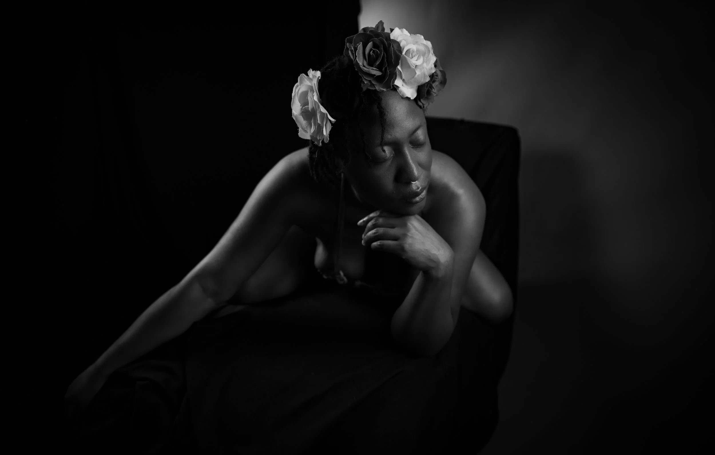 Black and white studio portrait of a woman in a floral rose crown, reclining pensively on a dark chair with dramatic low-key lighting.
