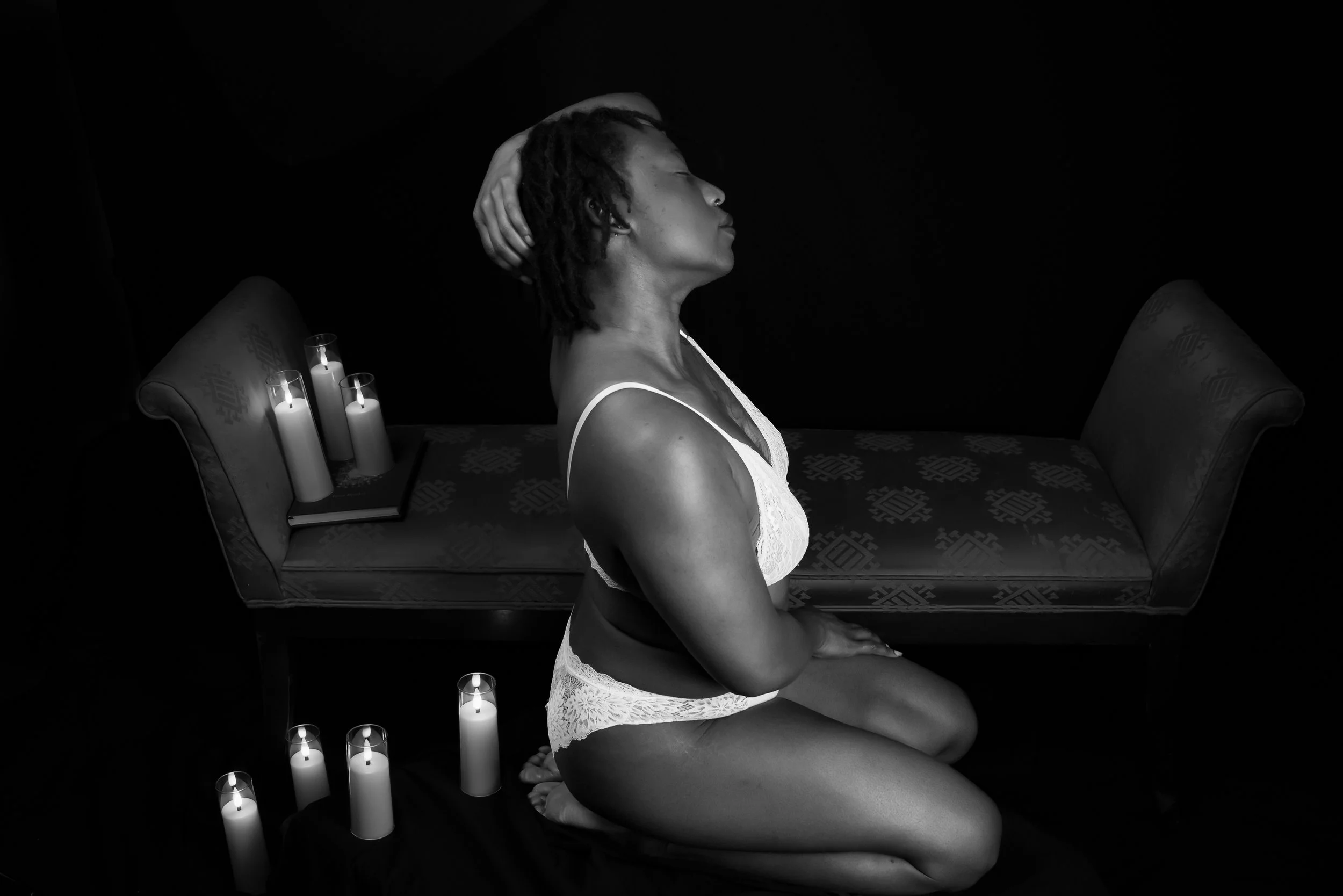A woman kneeling on a dark surface, surrounded by lit candles, with her eyes closed, head tilted back, and hand resting on her head, in front of a vintage-style chaise lounge.