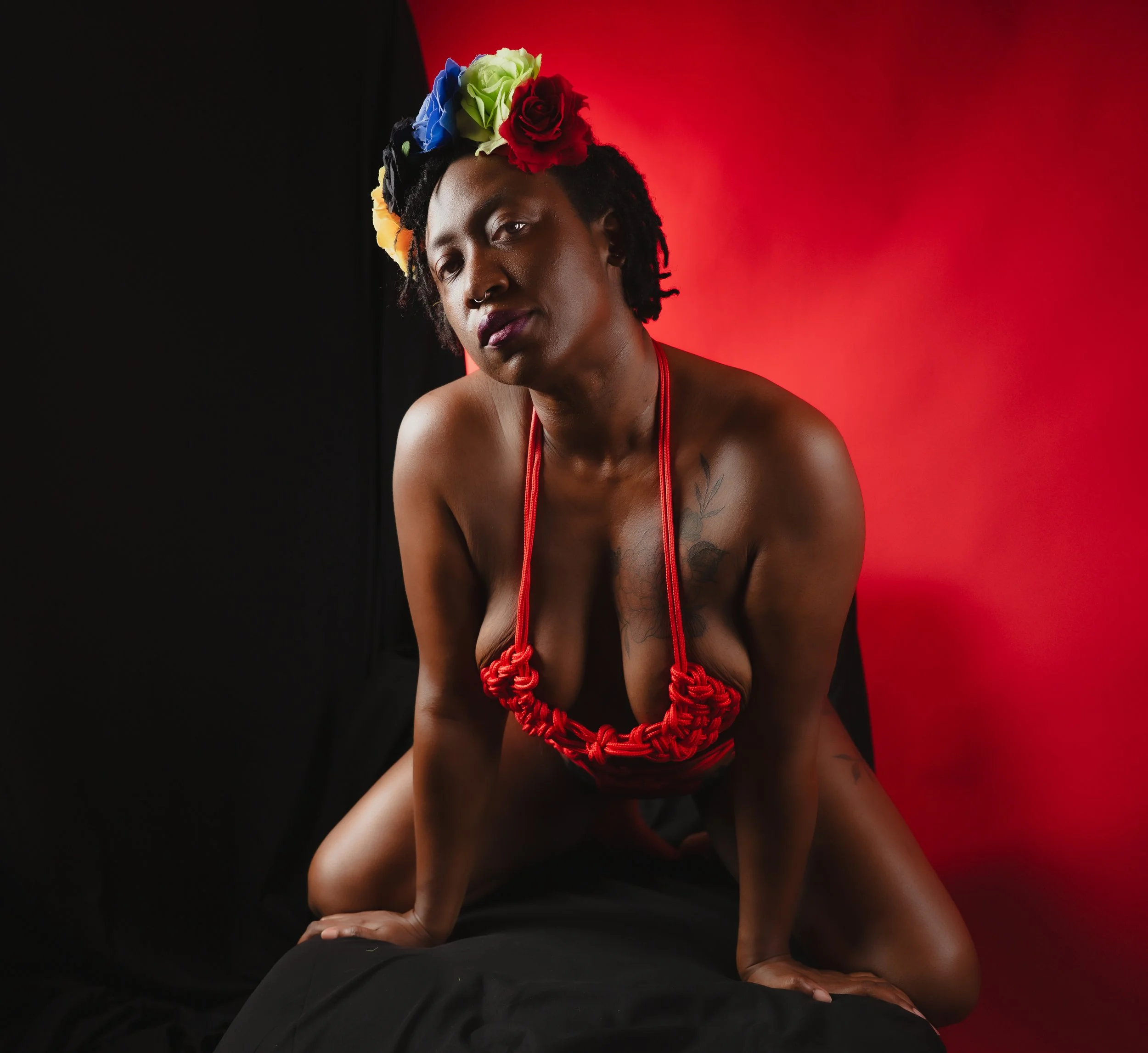 Bold shibari portrait of a woman in red rope harness and vibrant multicolor floral crown, kneeling against a split black and red background with a direct gaze.