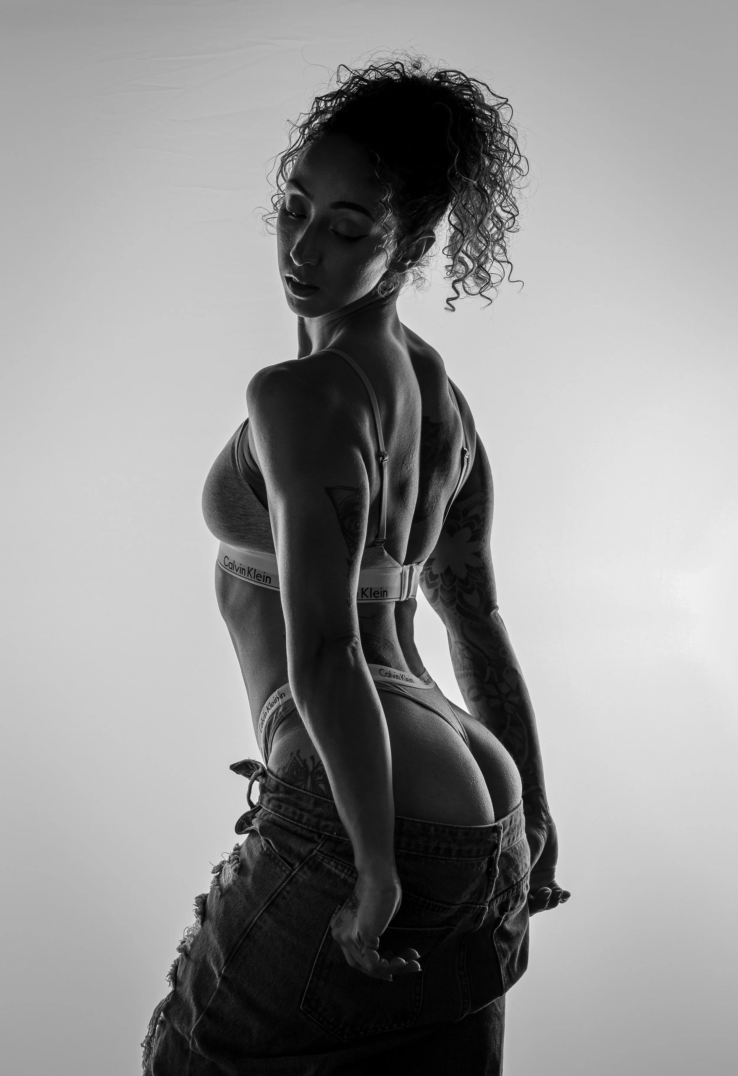 Black and white fitness portrait of a muscular woman shot from behind, showcasing defined back muscles with dramatic side lighting against a light gray background.