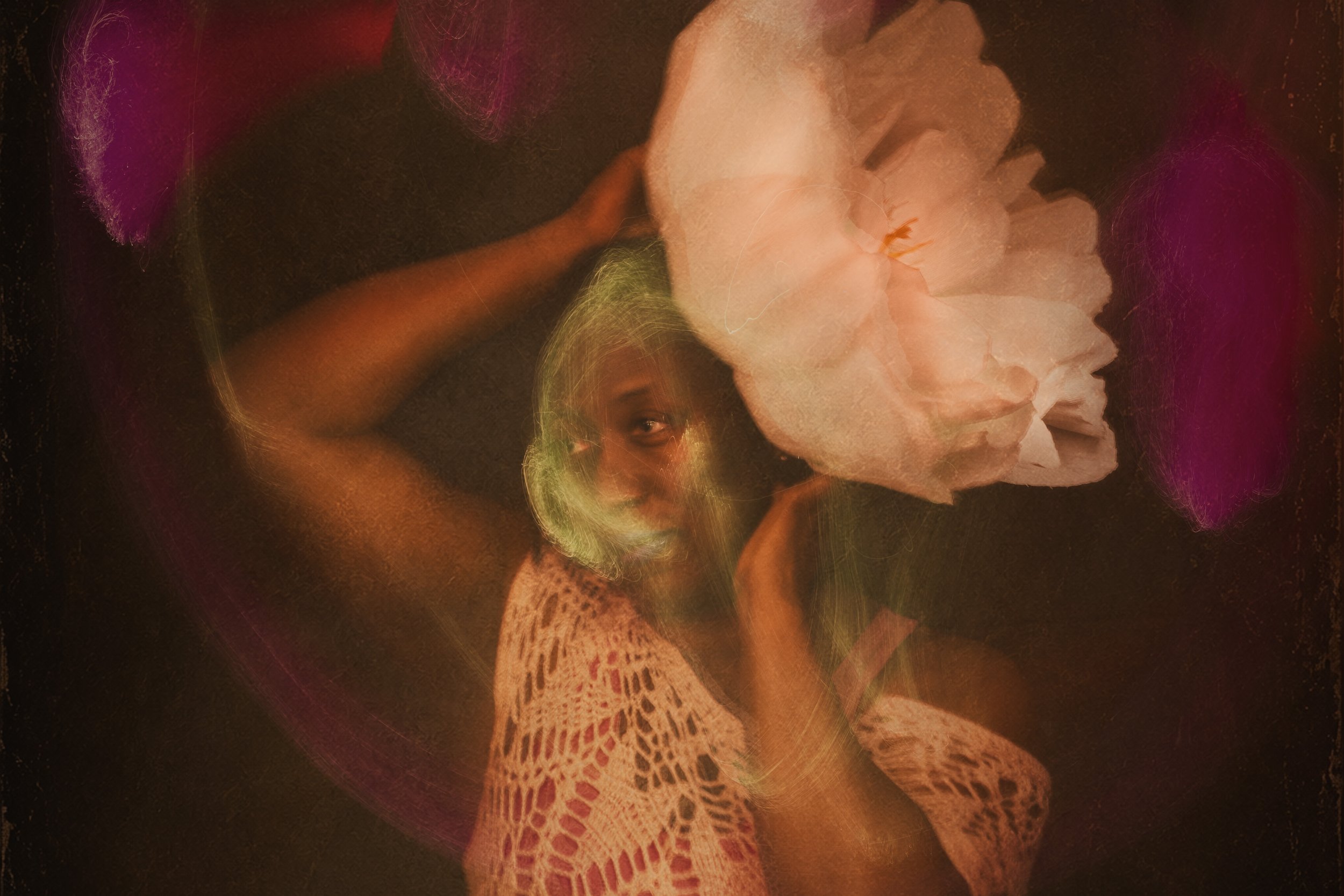 A woman with dark curly hair wearing a pink crocheted top posing with her eyes closed and head tilted back, surrounded by pink and gold light effects.  A large white flower is on her head.  Her face is lit by green light.  Light painting.