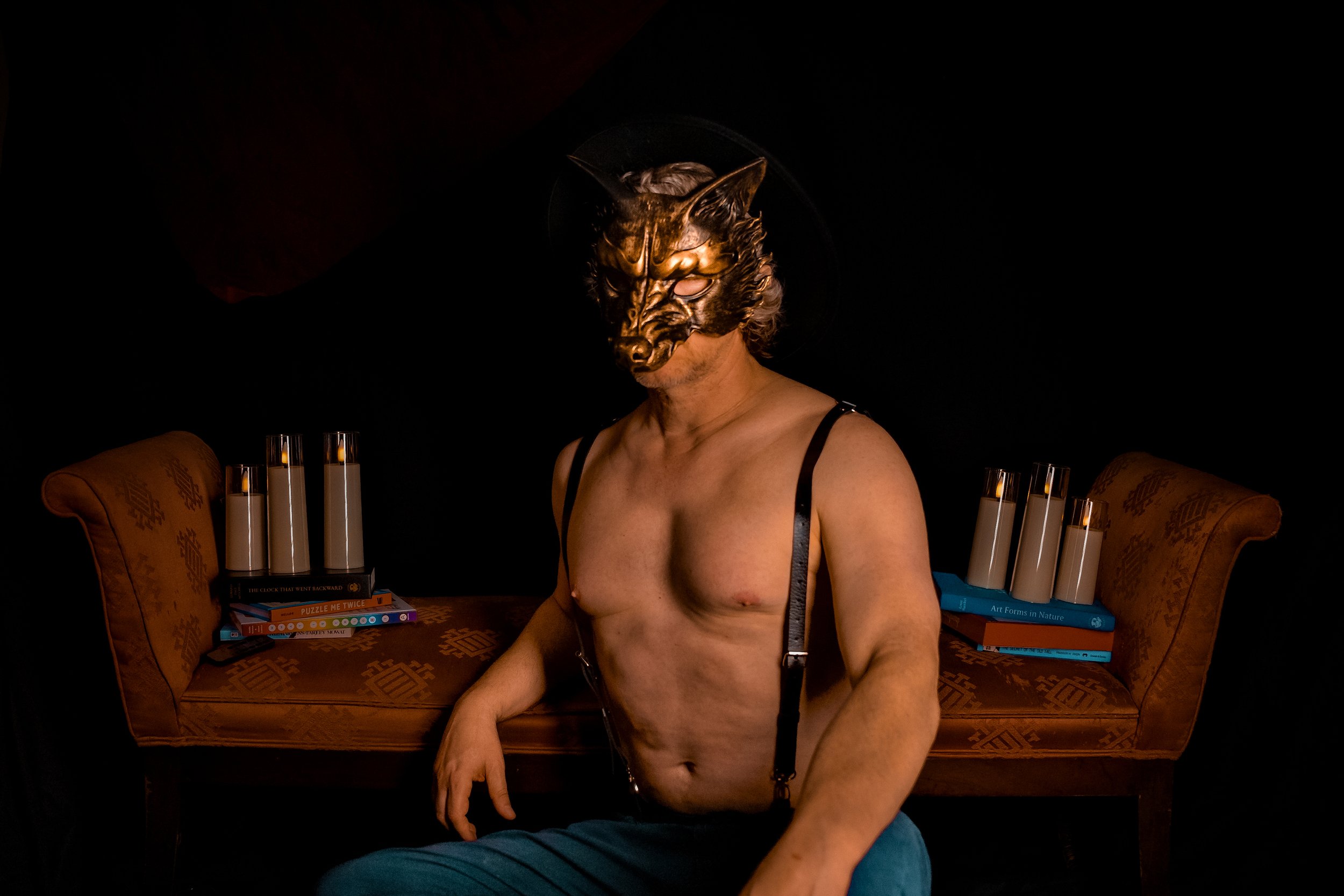 A shirtless man wearing a gold and black wolf mask, sitting on a chair in front of a dark background. There are stacks of books and glasses of milk on both sides of him.
