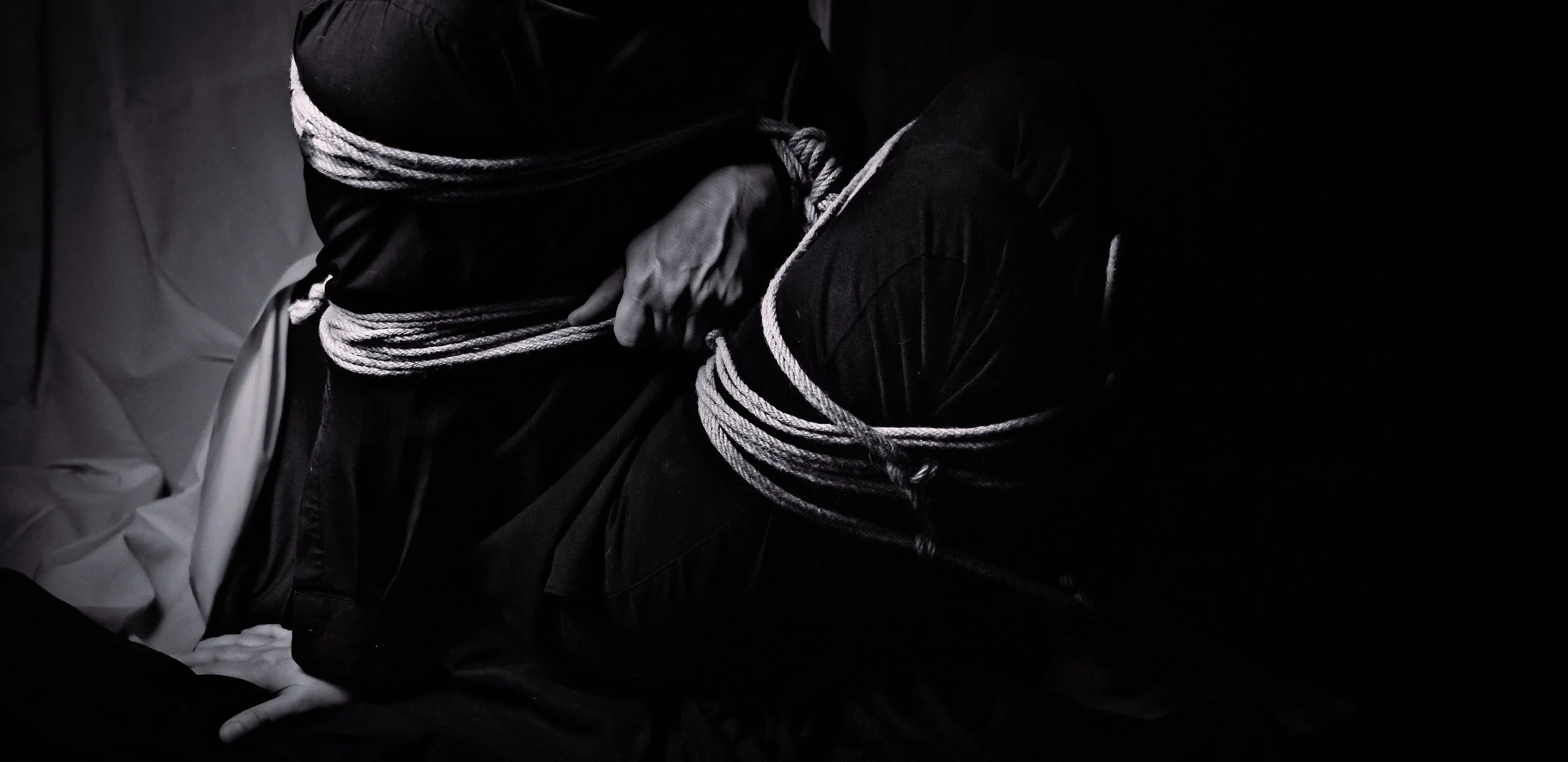 A person bound with white rope, sitting in a dark room with their face covered.