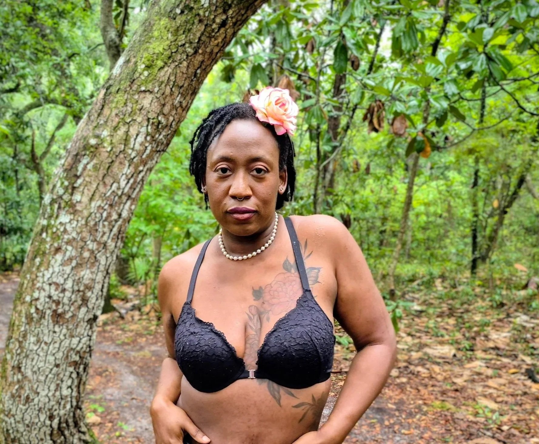 A woman with short dreadlocks, wearing a black lace bra, a pearl necklace, and a pink flower in her hair, standing outdoors among trees and greenery.