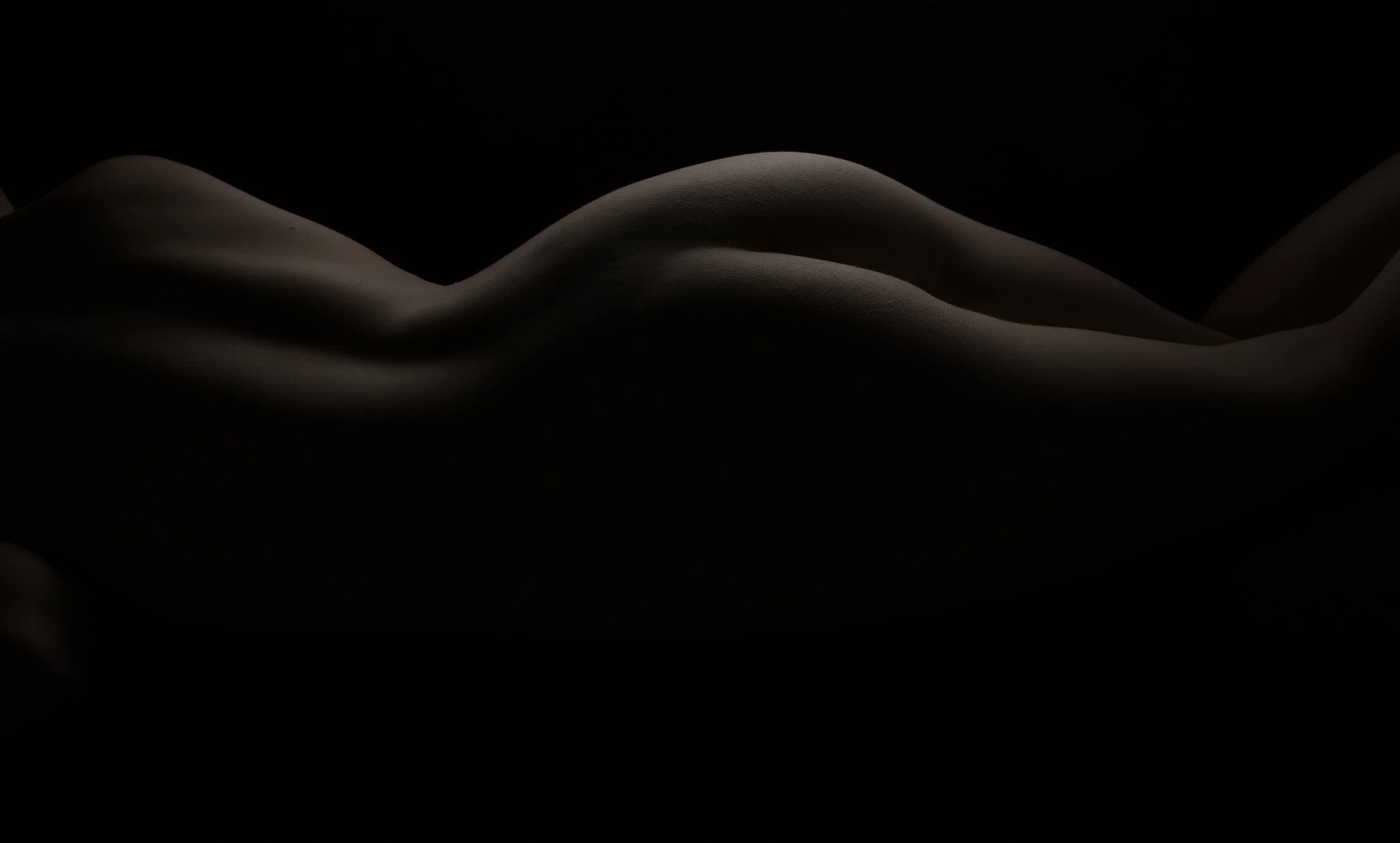 Close-up of a human body lying on a dark background, illuminated softly to highlight the curves and contours of the skin.