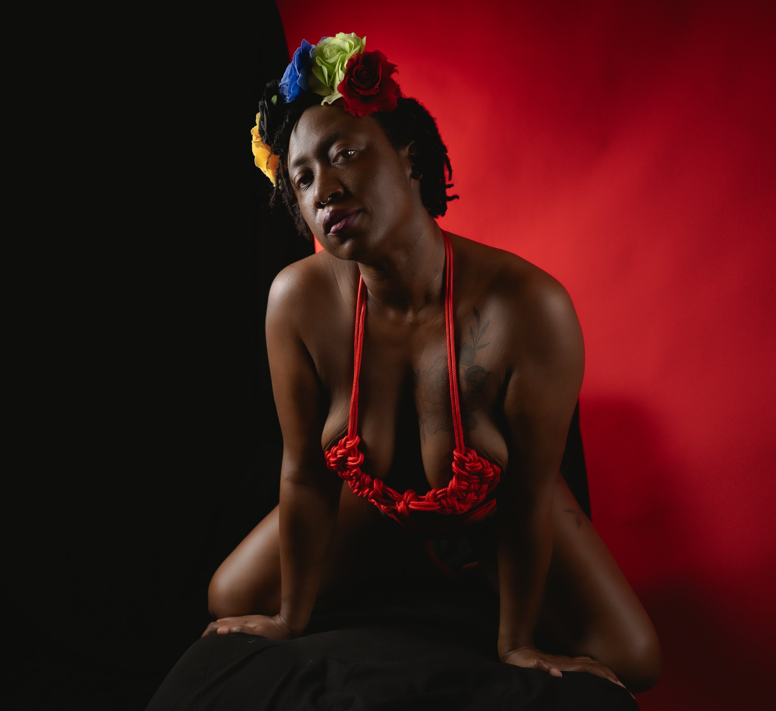 A woman with dark skin and dreadlocks wearing a floral headdress, a red necklace, and knot rope accessories, posing against a red and black background.
