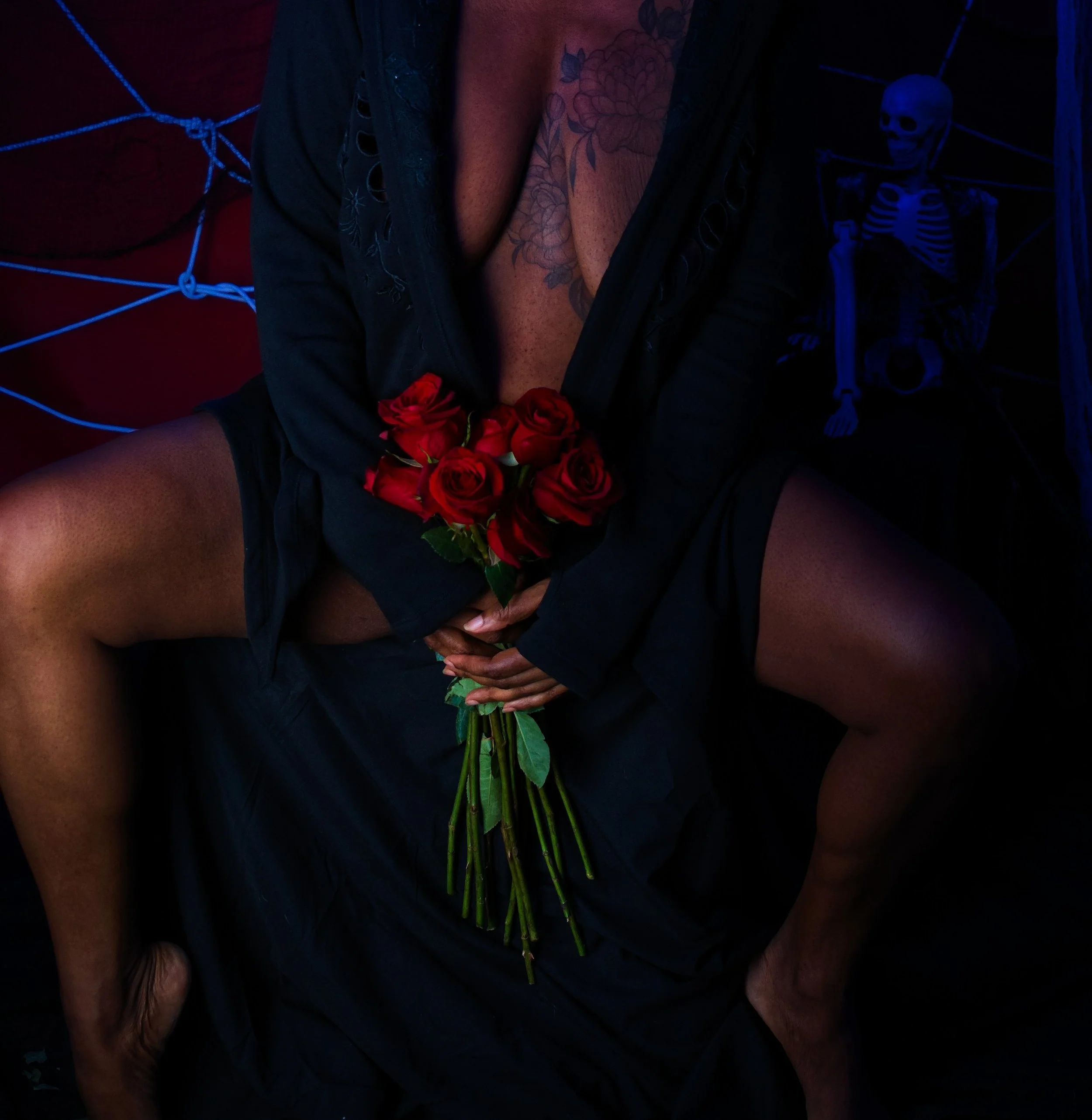 Person sitting in a dark room holding a bouquet of red roses, with tattoos visible on their chest and neck, and a skeleton decoration behind them.