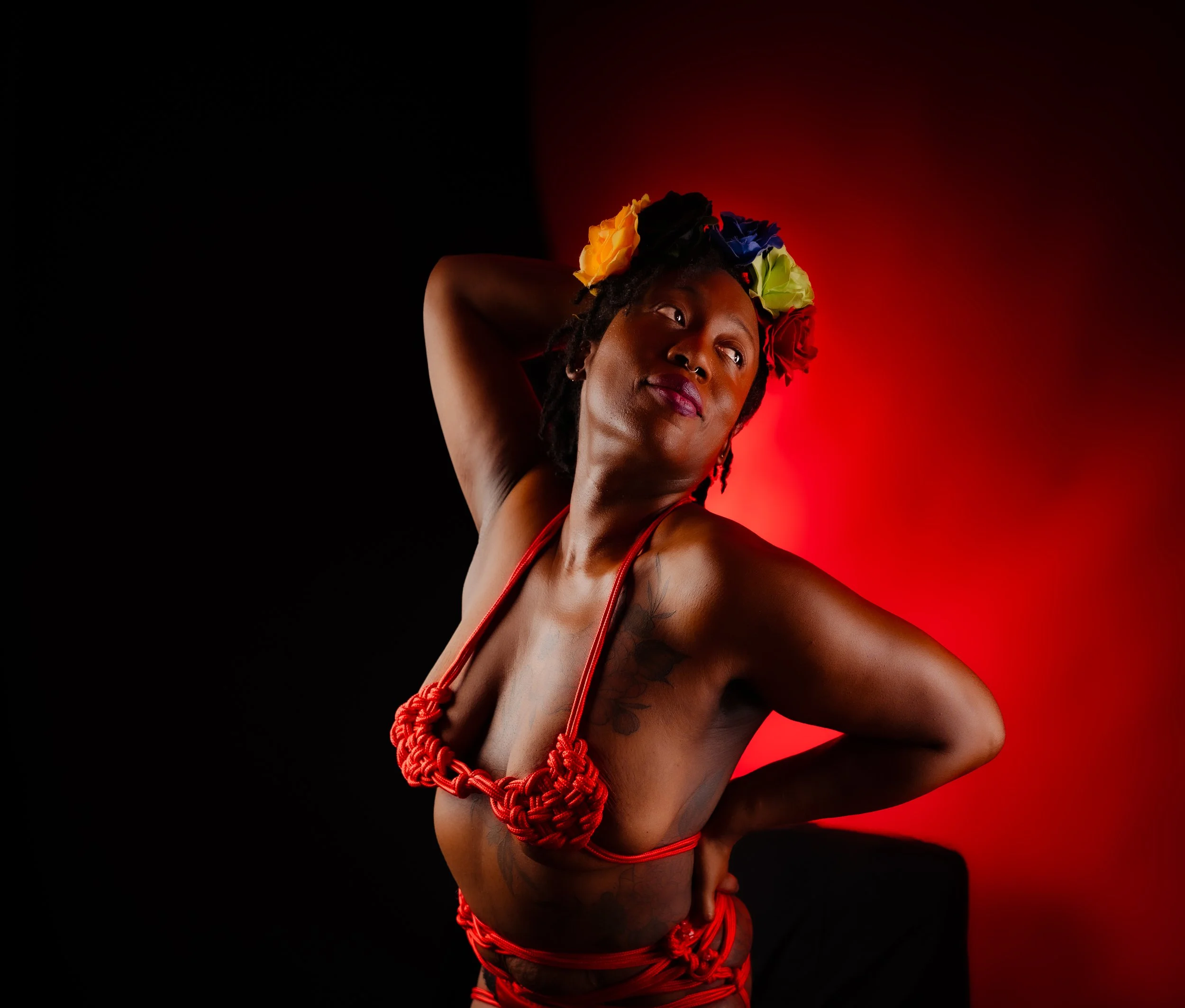 Vibrant studio portrait of a woman tied in red rope with a colorful floral hair accessory, posed confidently against a split black and red background.