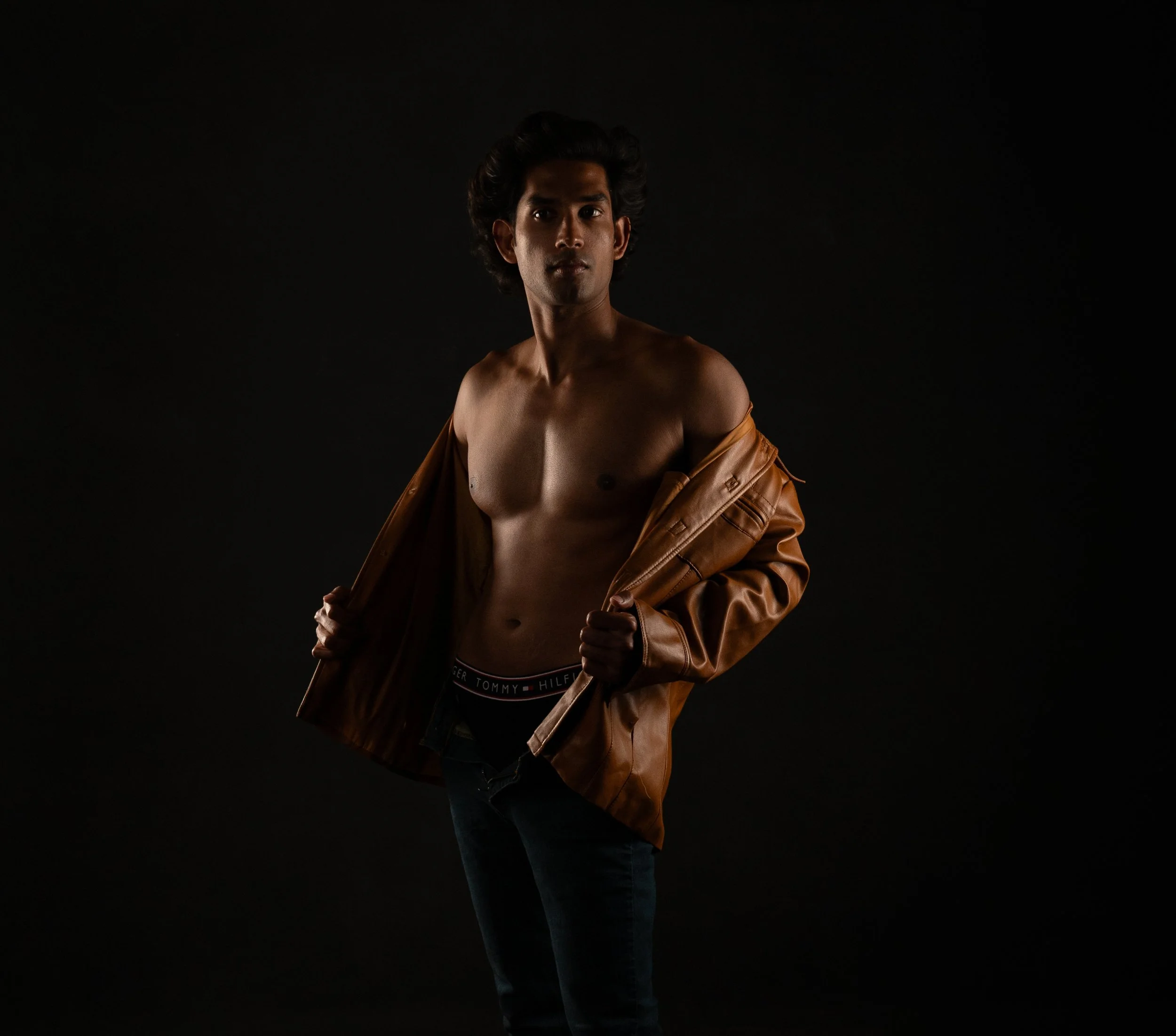 Low-key studio portrait of a shirtless man opening a tan leather jacket, lit with warm directional light against a dark background.