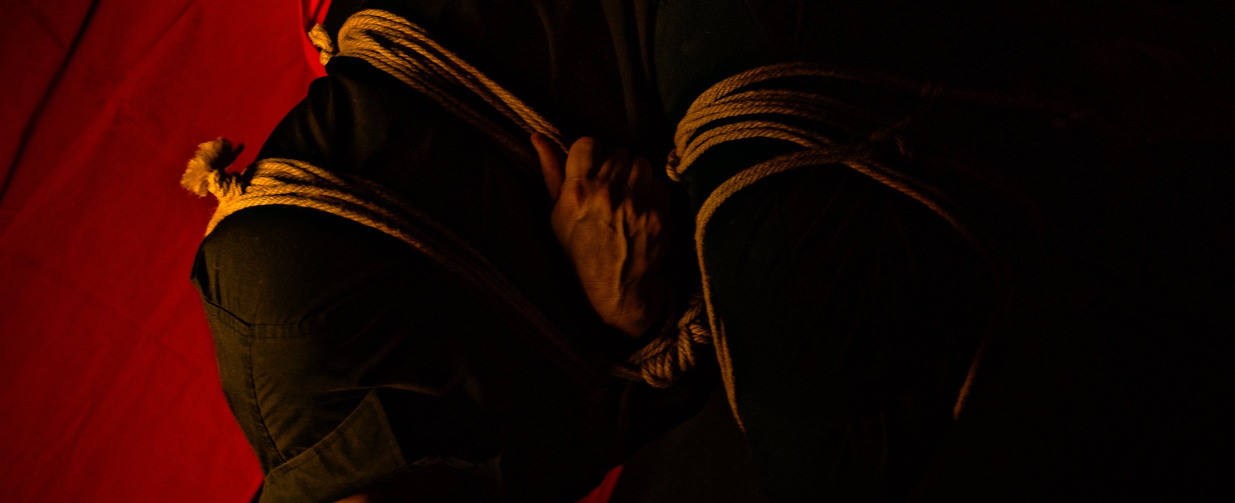 A person with a dark jacket and pants sitting on a red surface, with hands clasped together and a thick rope wrapped around their shoulders.