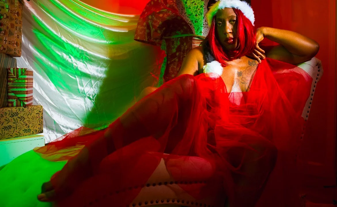 A woman wearing a Christmas Santa hat and red tulle dress, sitting on a chair, surrounded by Christmas presents and holiday decorations, with colorful lighting.