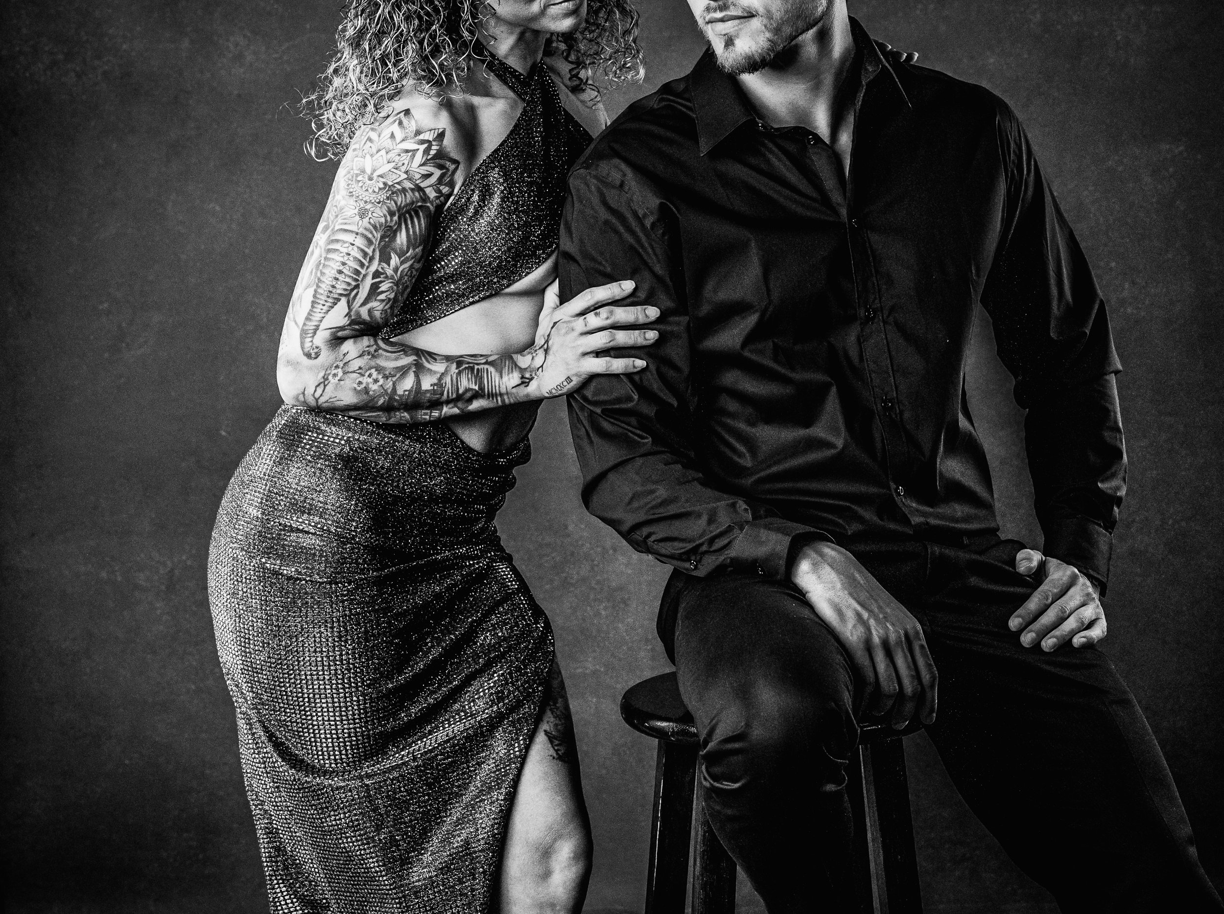 Couples Photography.  Black and white photo of a woman with curly hair and tattoos, and a man in a black shirt and pants, standing close together, with the woman resting her hand on the man's arm.