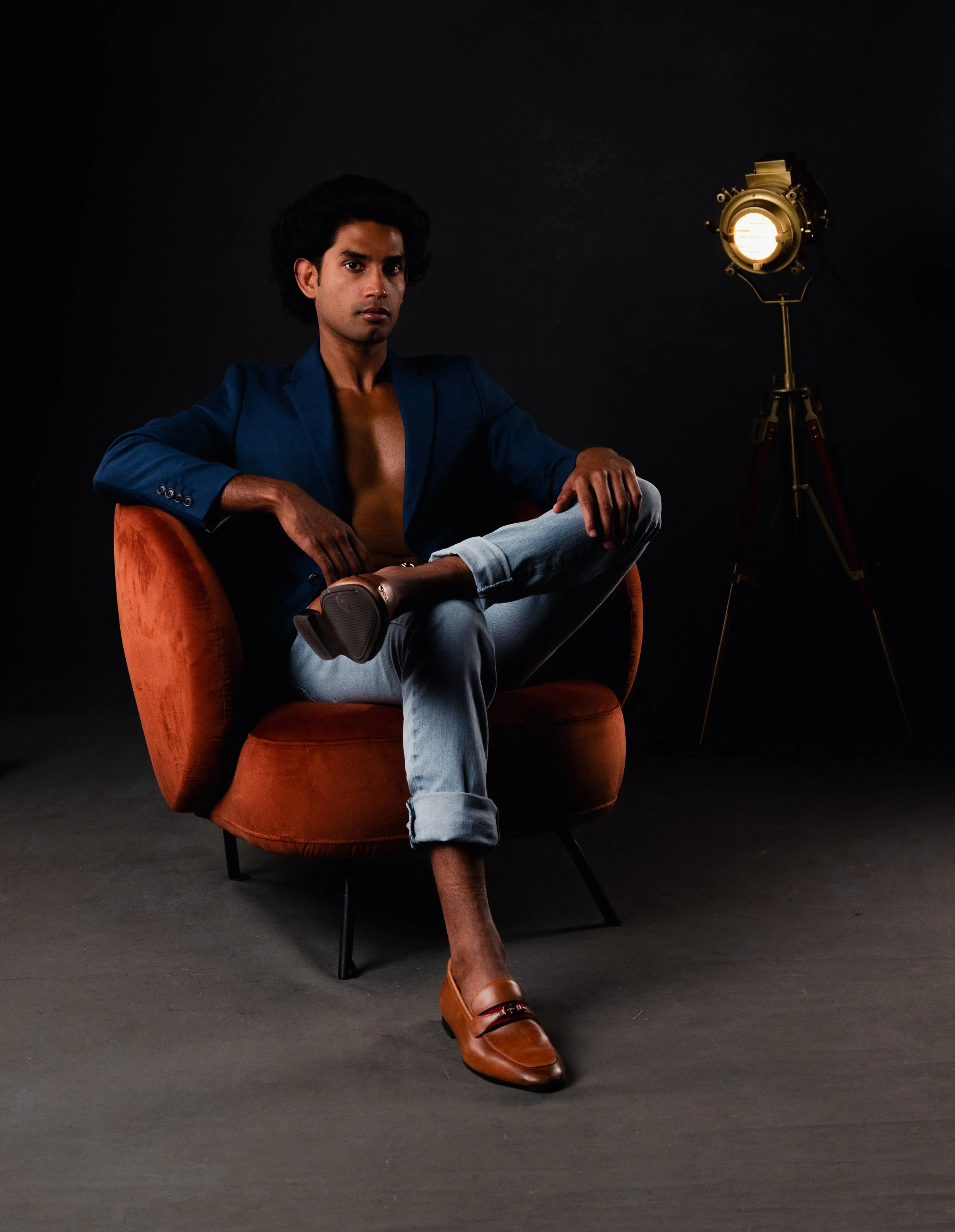 A man sitting on an orange cushioned chair in a dark studio setting, wearing a partially open blue blazer, light blue jeans, brown shoes, and a ring, with a professional studio light on a tripod stand to his right.