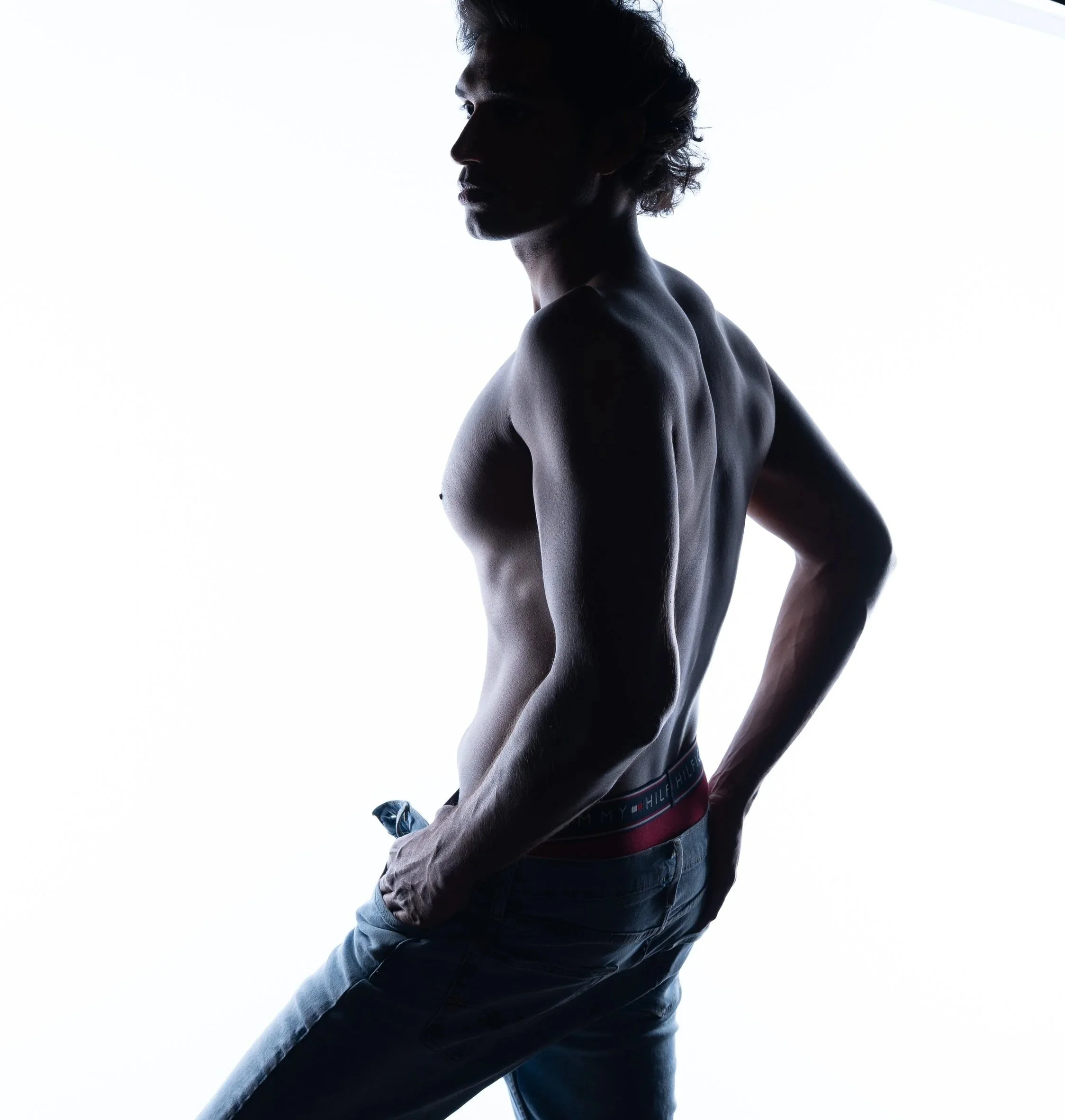 Silhouette of a shirtless young man wearing jeans, standing against a white background with his left arm bent and hand near his hip, looking to the side.