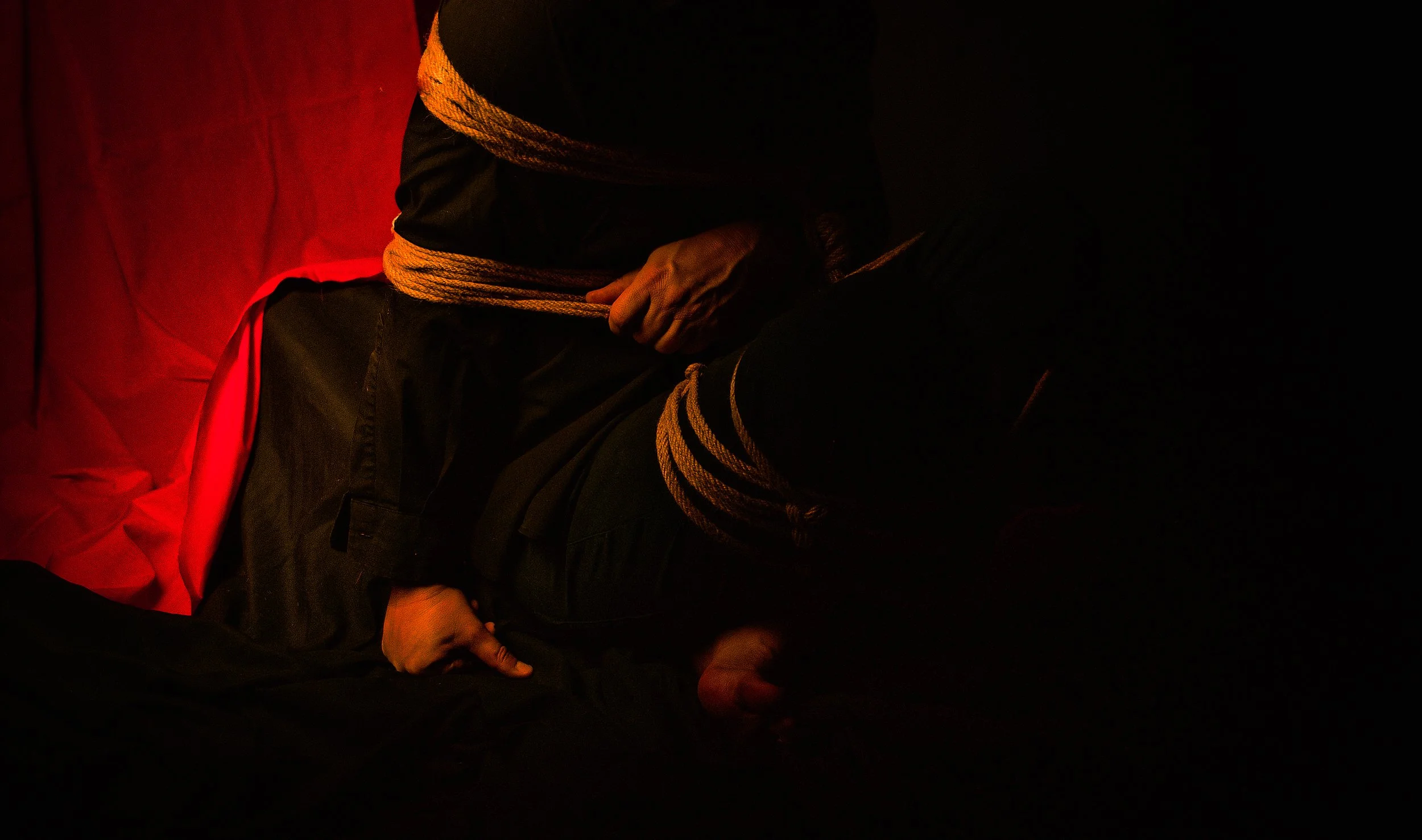 Person with hands bound sitting on a red and black surface in low light.