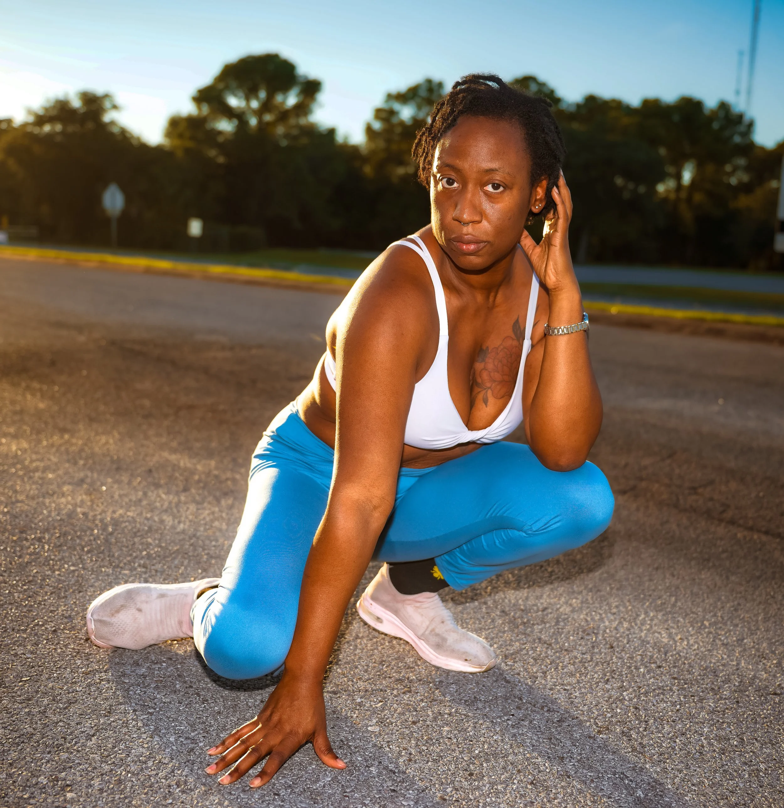 A woman with short dreadlocks, wearing a white sports bra, blue leggings, and white sneakers, squatting on a paved road during sunset, with trees and power lines in the background.