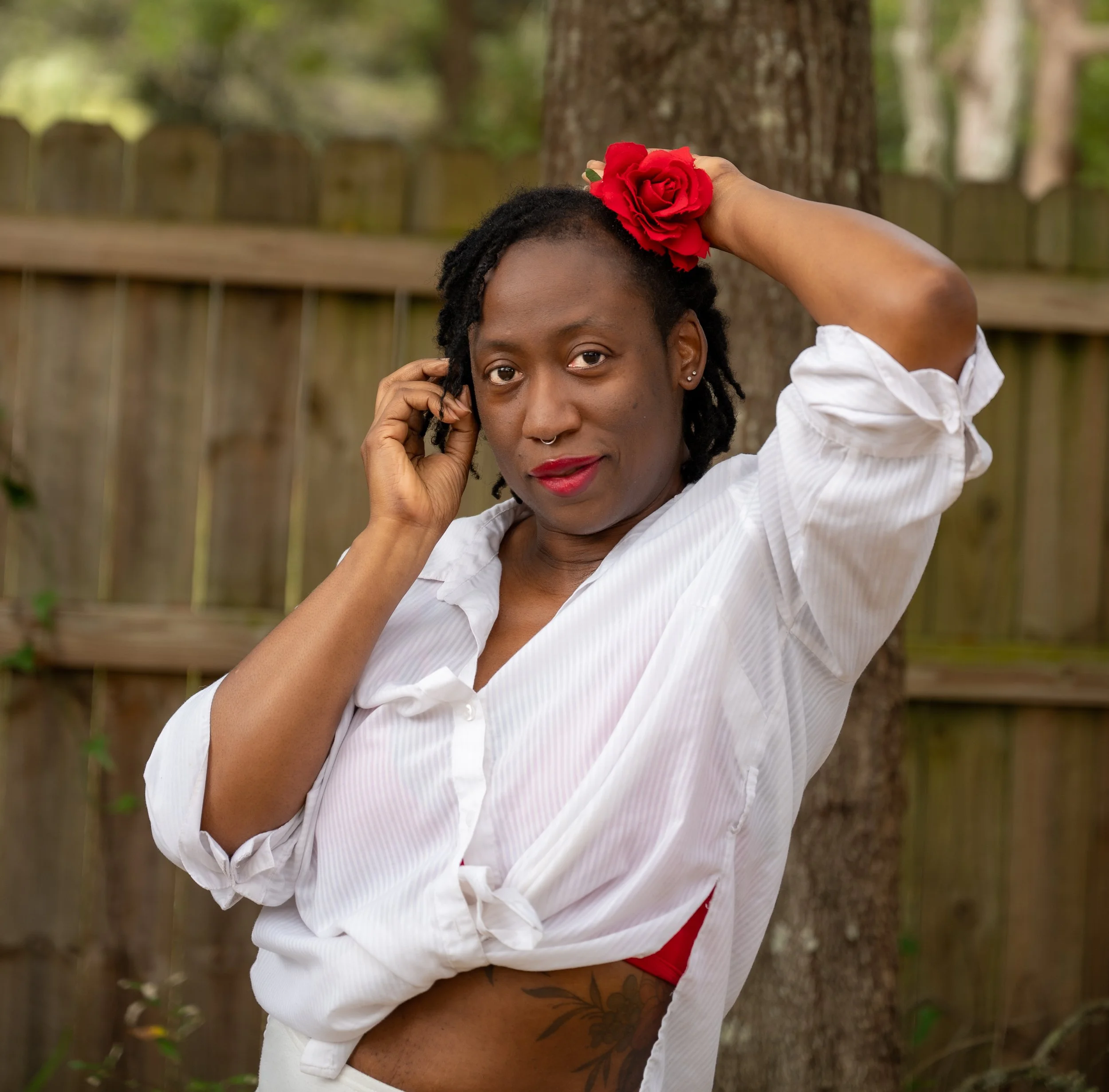 A woman with dark curly hair, wearing a white shirt, holding a red flower in her hair, outside near a tree and wooden fence, looking at the camera.