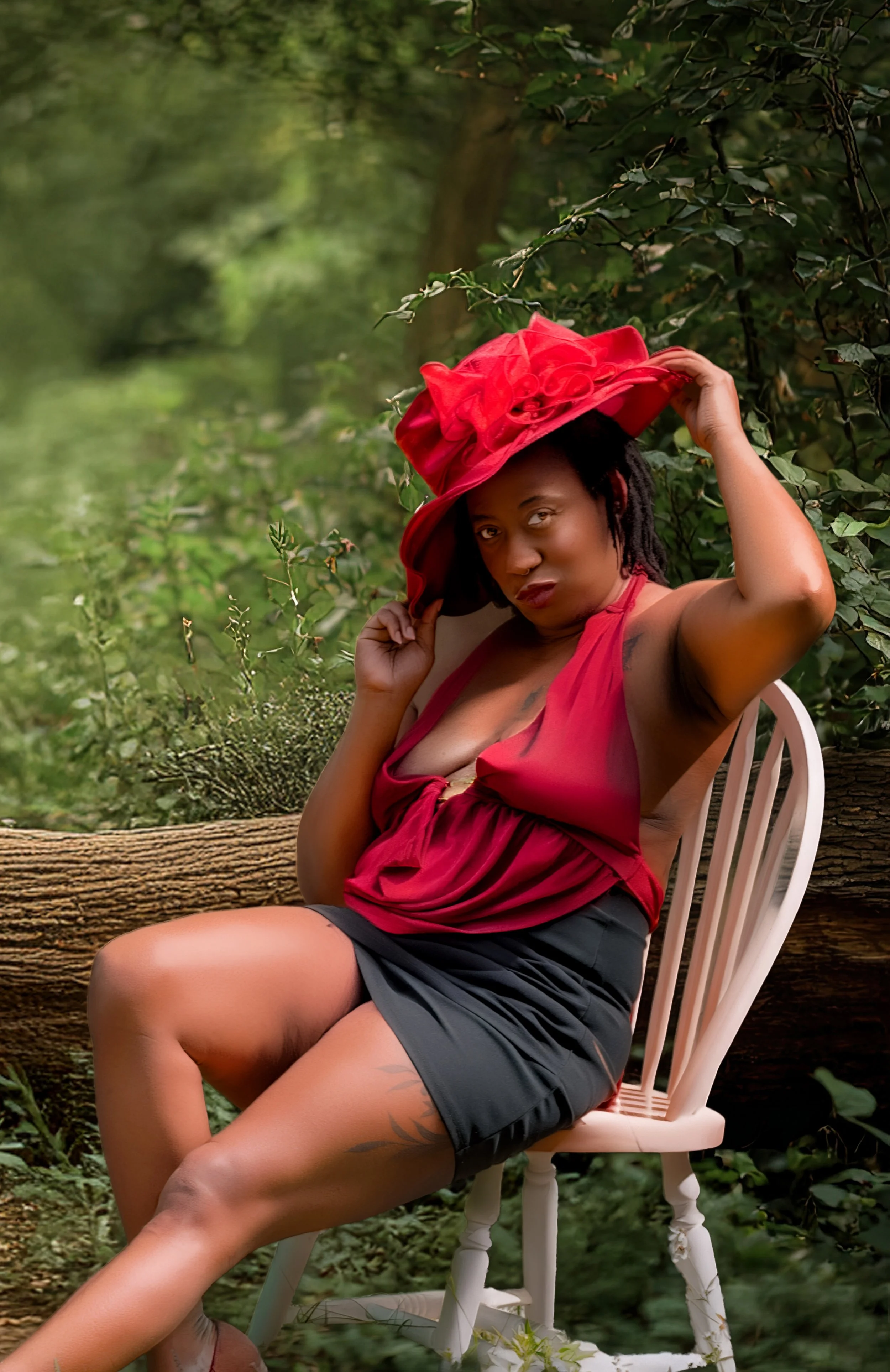 A woman with dark skin and dreadlocks sitting on a white chair in a lush outdoor setting with green foliage, wearing a red sleeveless top, black shorts, and a large red hat, with her hand holding the hat.