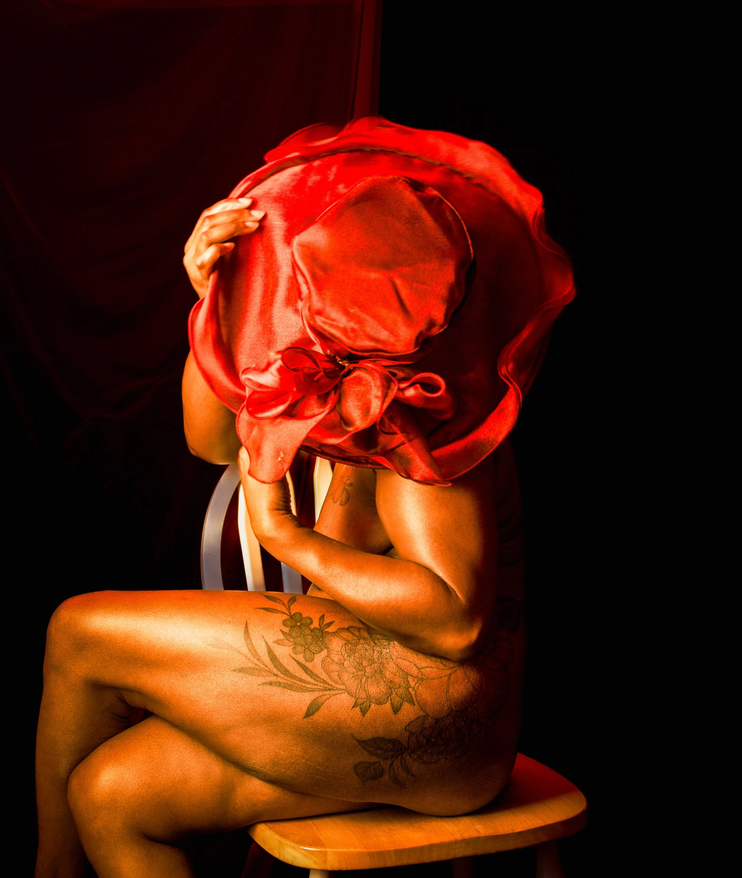 Person sitting on a stool, partially nude, with a large red satin hat covering their face and head.