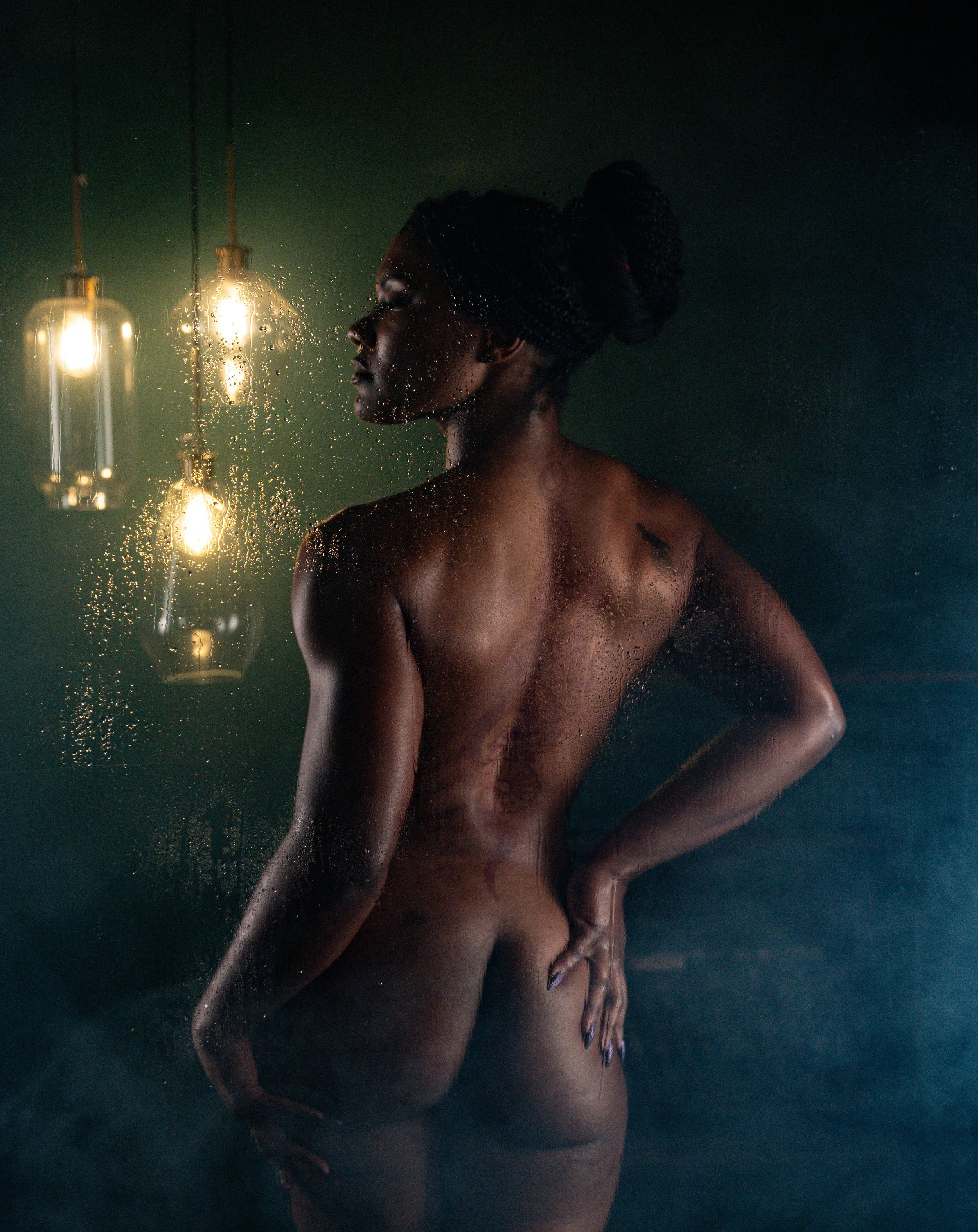 Fine art nude of a woman shot from behind, hands on hips, with warm pendant lighting and atmospheric mist against a dark teal background.