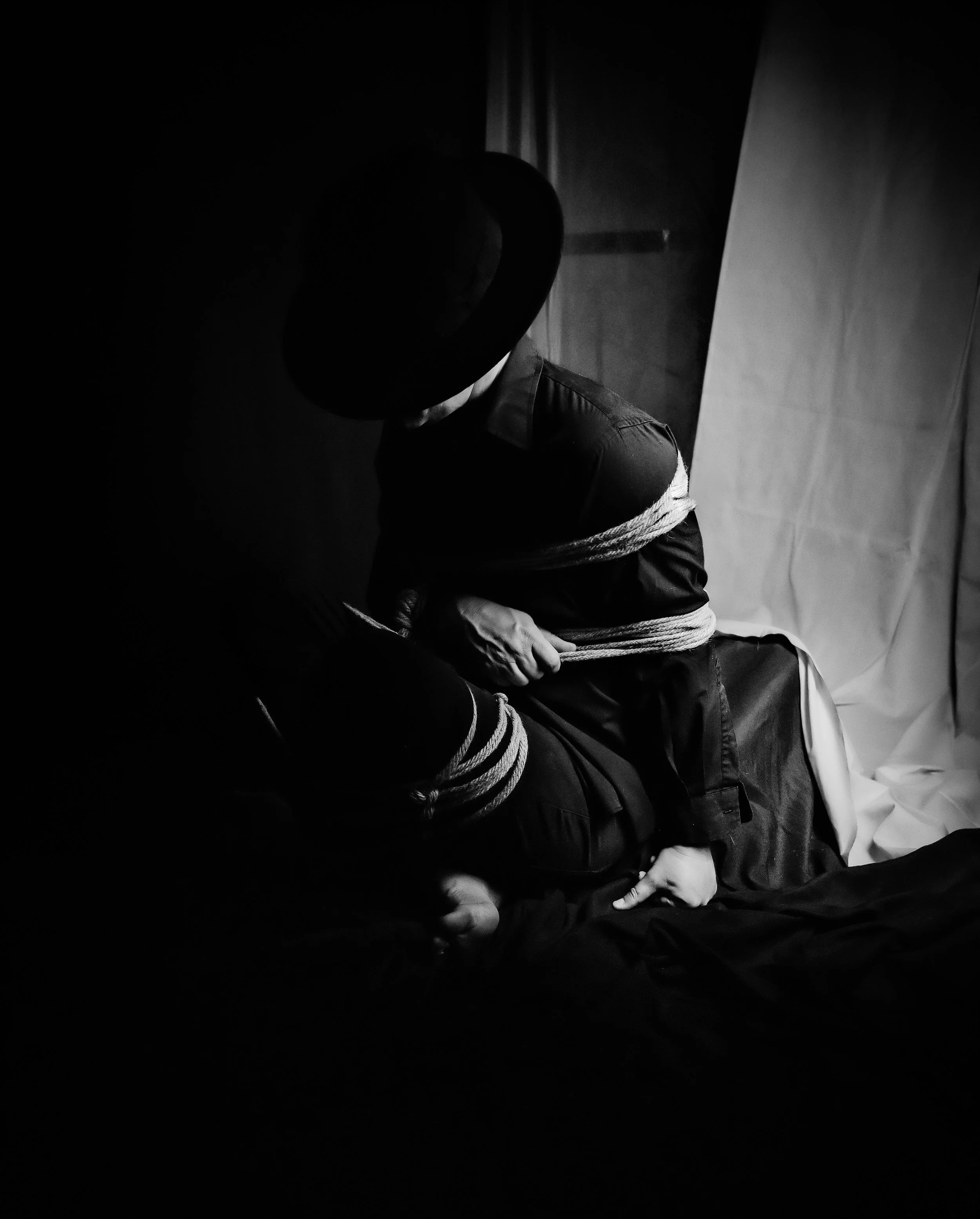 A person wearing a hat and dark clothing is tied up with ropes and sitting on the floor in a dark room.