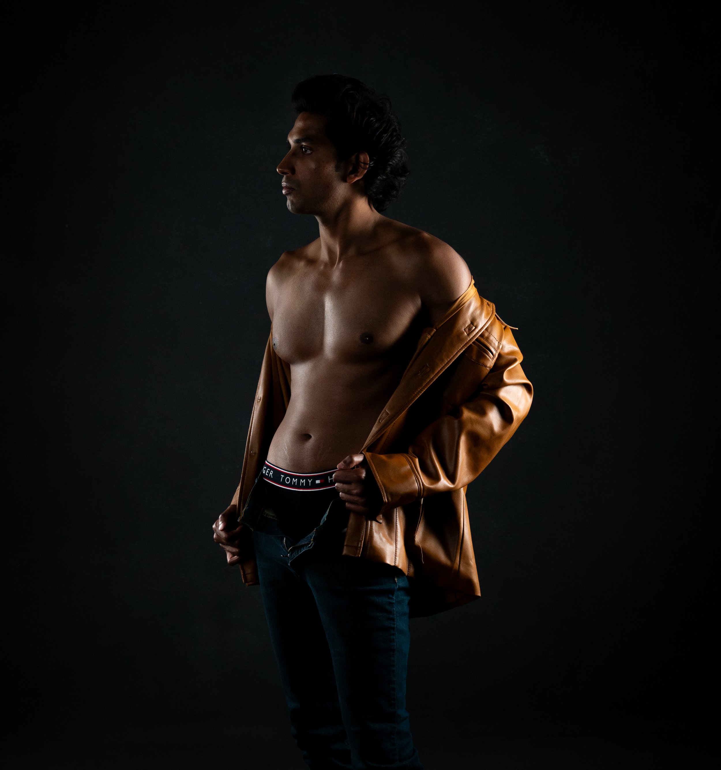 Moody studio portrait of a shirtless man in jeans wearing an open tan leather jacket, lit with warm side lighting against a dark background.  Boudoir for Men.  Male Boudoir. Gentlemen Boudoir.  Dudeoir and masculine boudoir.