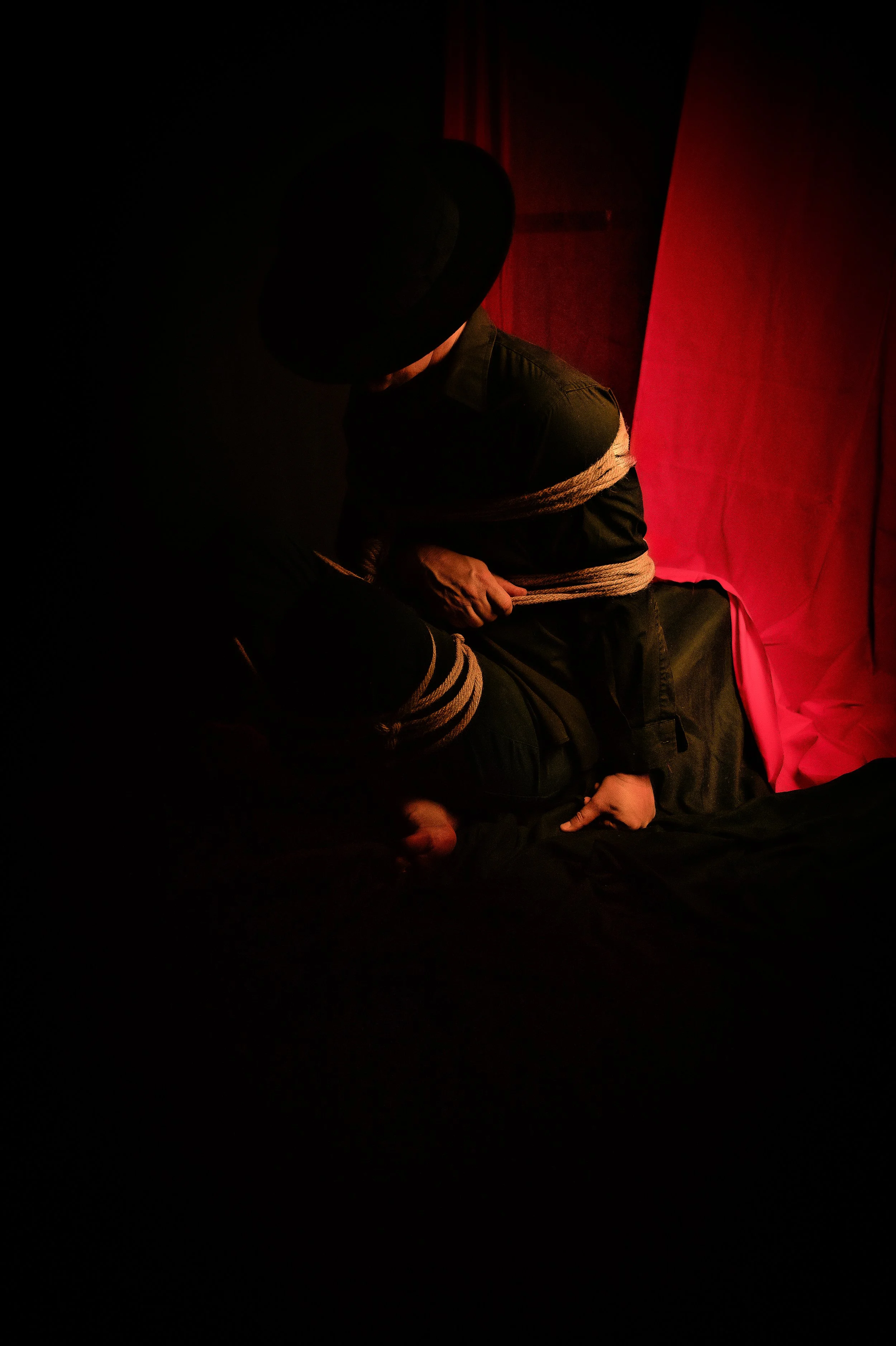 A person in dark clothing and a hat is tied up with ropes, sitting on the floor, with another person leaning over them in a dimly lit room with red curtains.