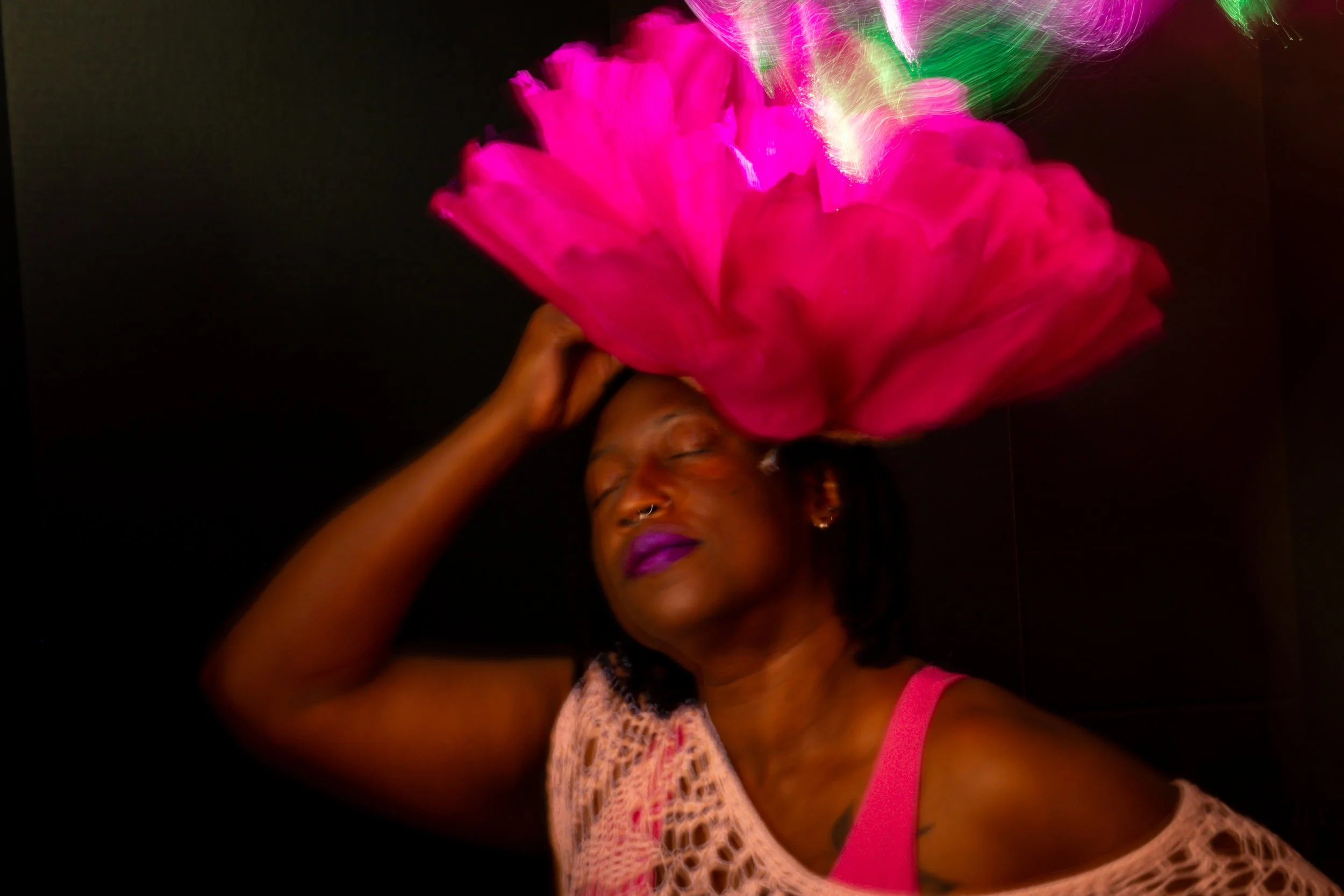 Painterly light painting portrait of a woman with eyes closed and purple lipstick, balancing a large vivid pink peony on her head while soft green and pink light trails swirl above, wearing a white pink top, Bellissimo Expressions studio.