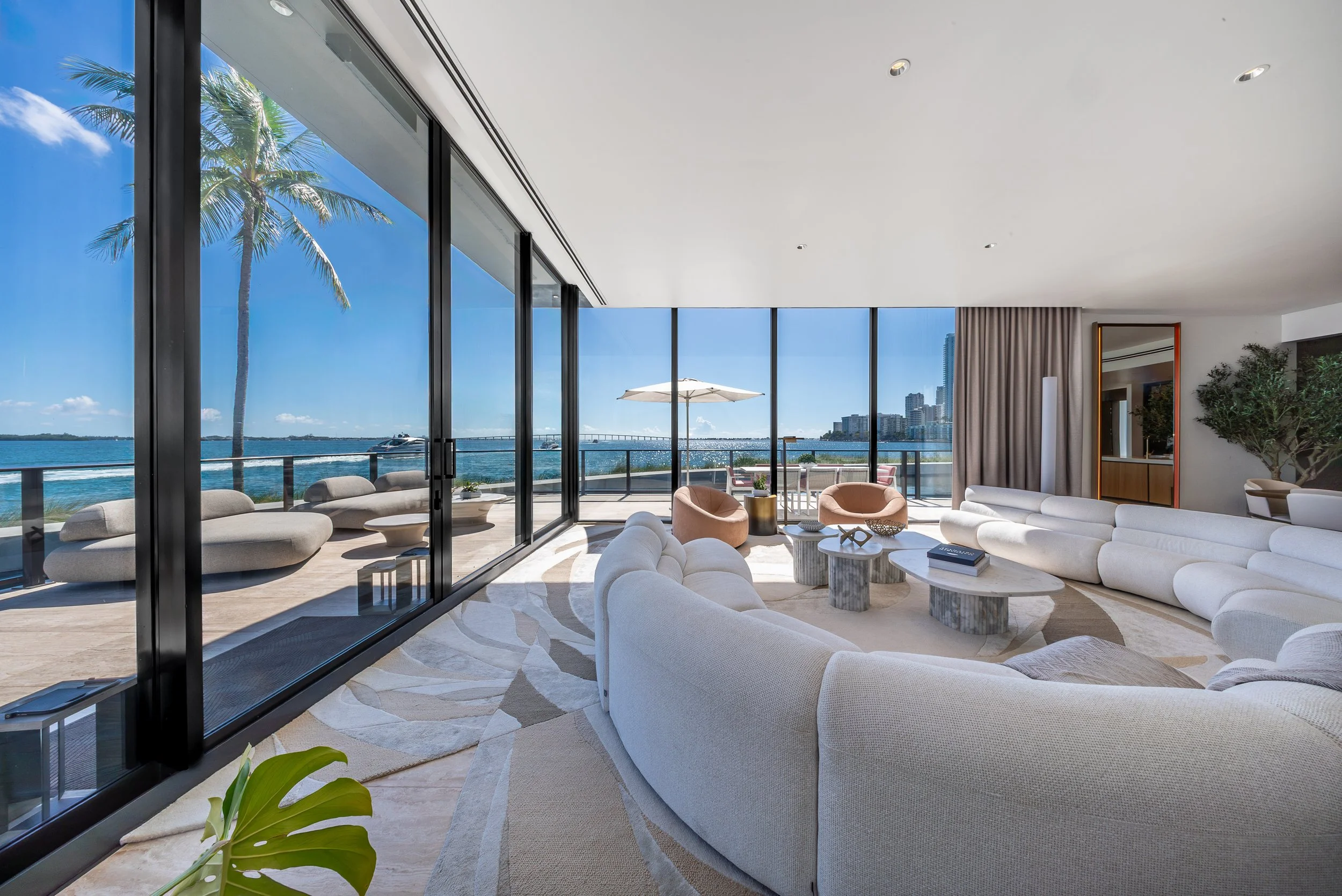 Modern living room with large glass windows showing ocean view, outdoor balcony with seating, and city skyline in the background.