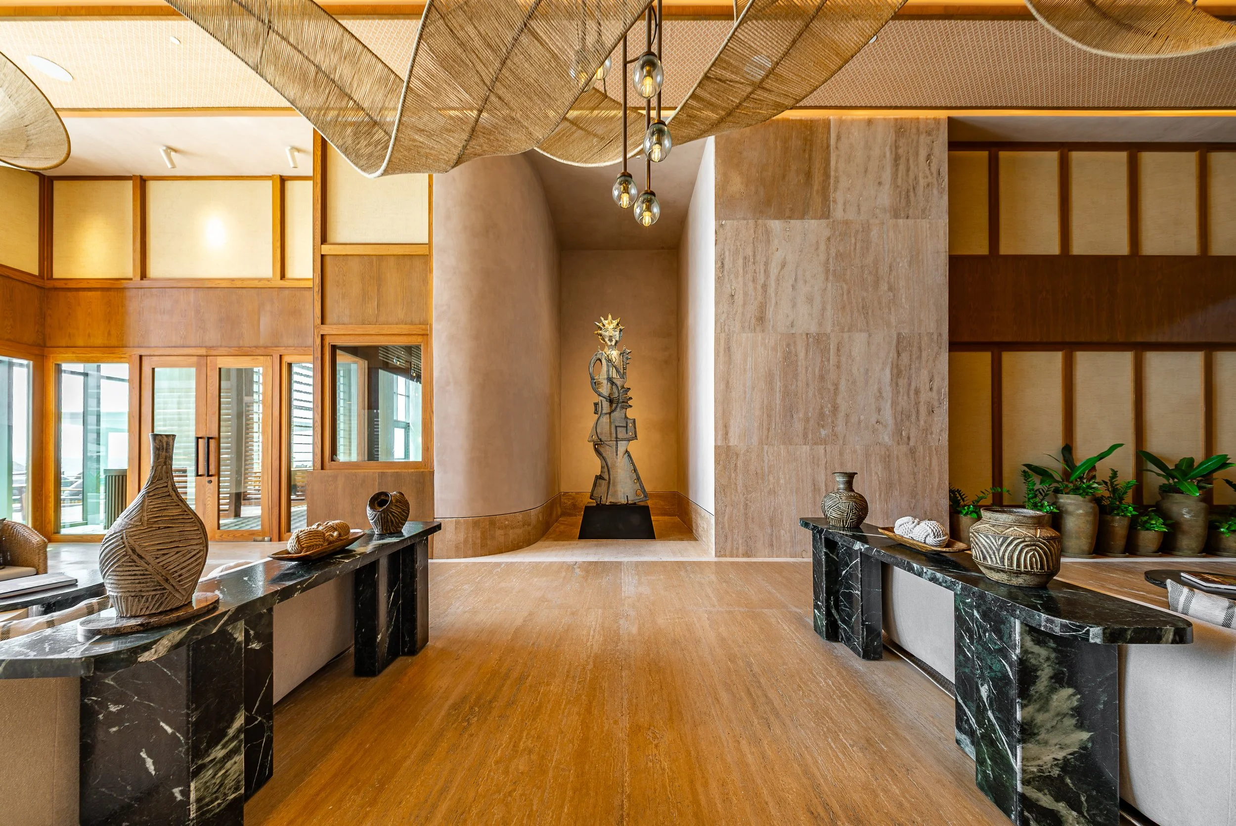 Elegant indoor space with wooden and stone decor, including pottery and sculptures, sunlight through large windows, and modern lighting fixtures.