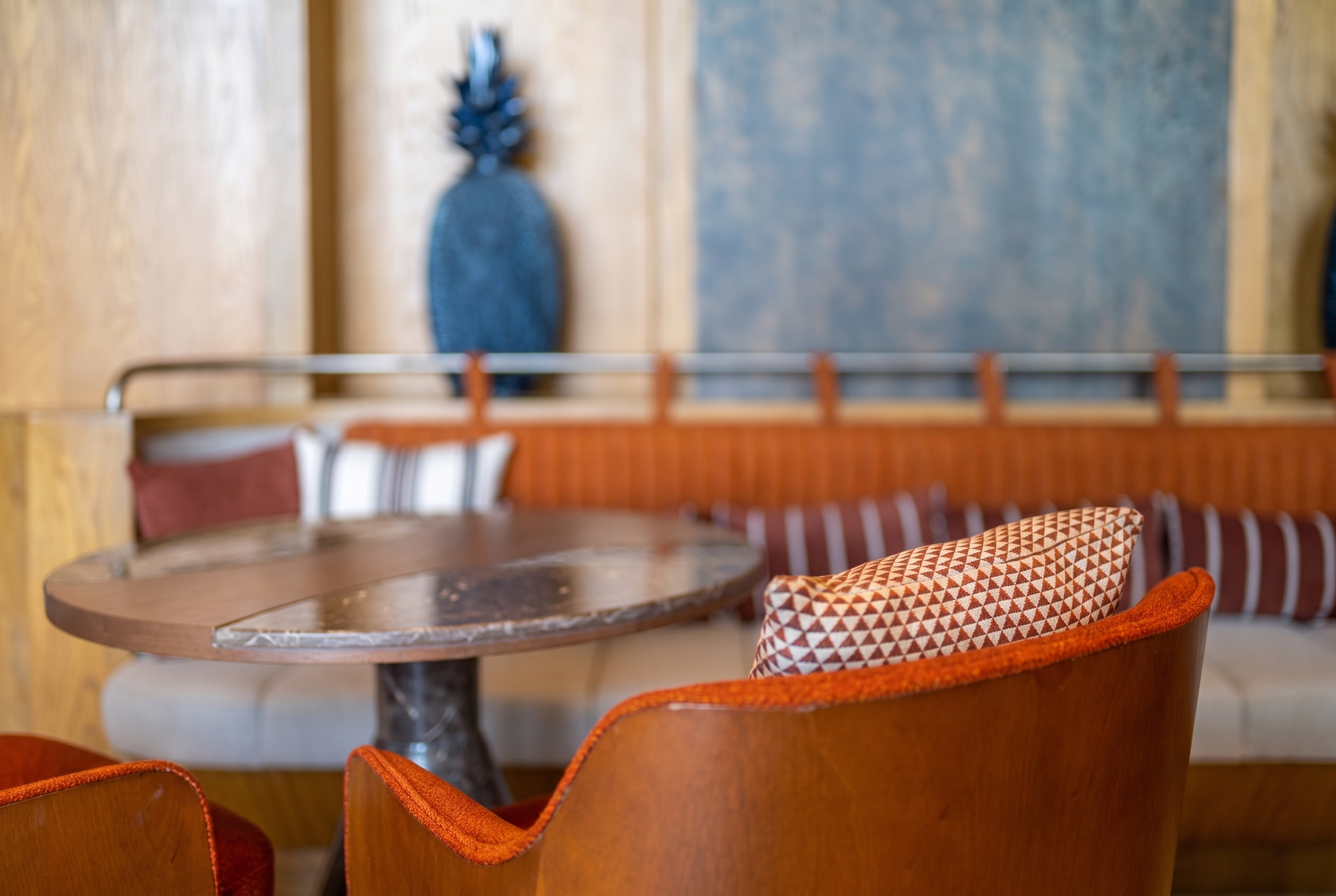 A cozy indoor scene with a small round wooden table, orange upholstered chairs with patterned fabric, a blue textured wall hanging in the background, and a wooden wall with modern decor.