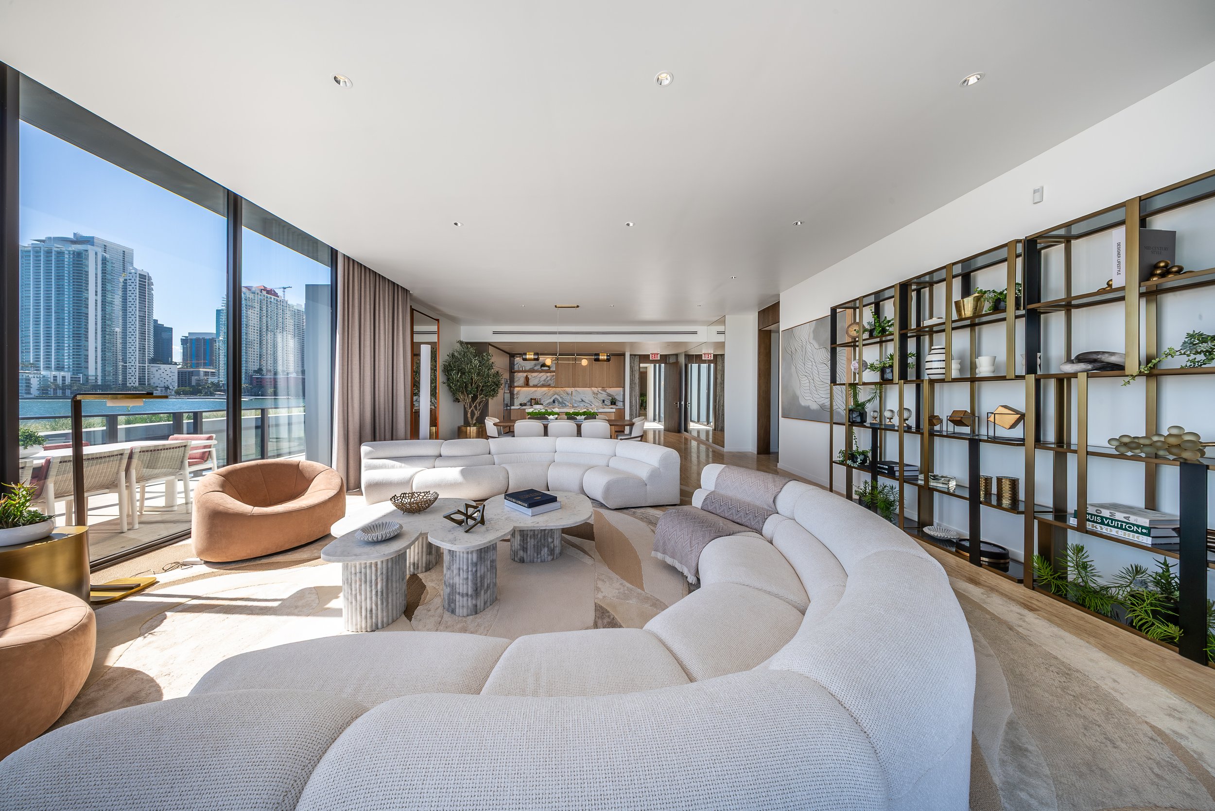 Spacious modern living room with large windows overlooking city skyscrapers, featuring curved white sofas, a round marble coffee table, a beige lounge chair, books, plants, and a decorative wall shelf.