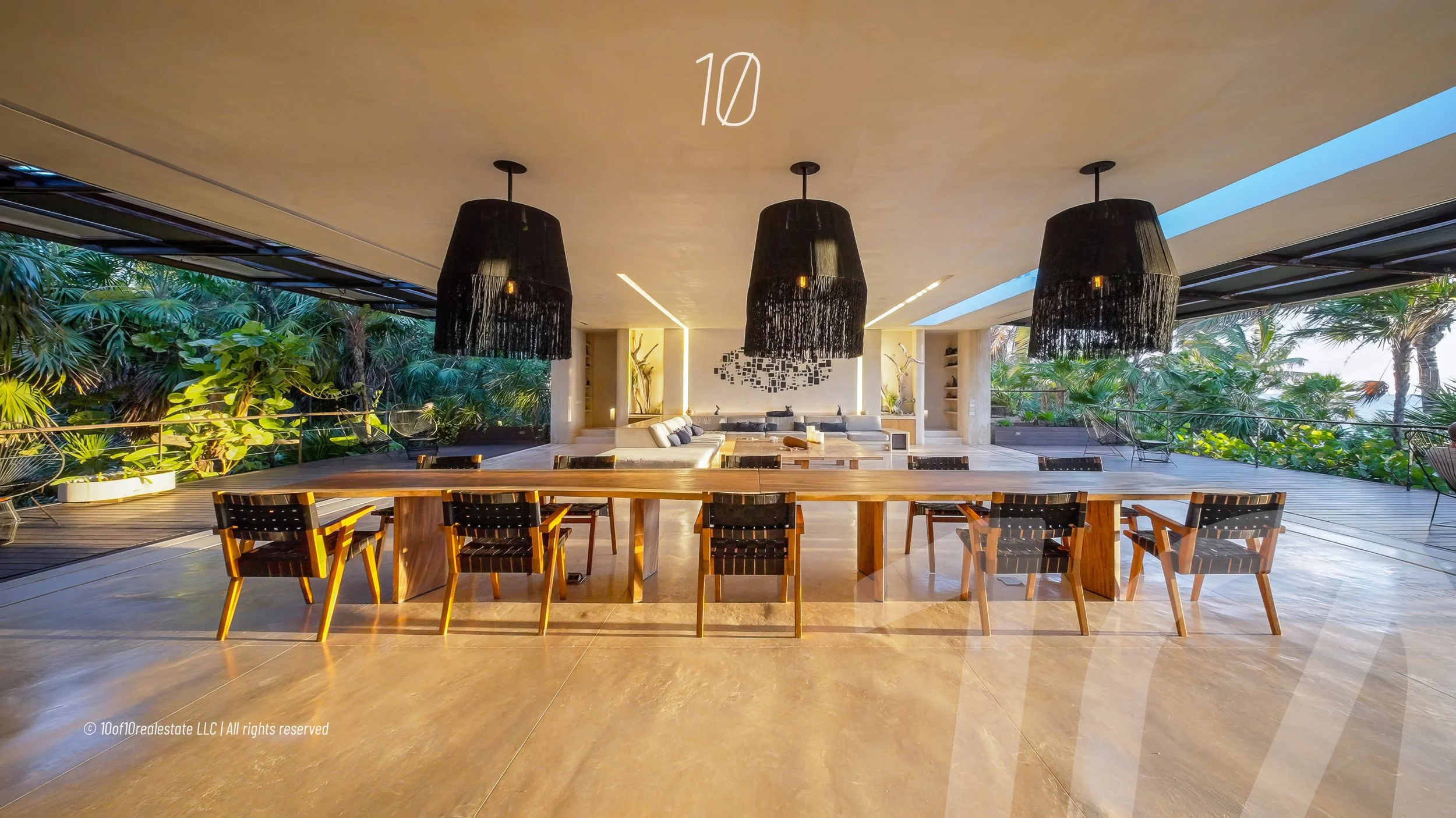 Modern open-concept living and dining area with large windows overlooking lush greenery, featuring a long wooden dining table, black pendant lights, and a sitting area with sofas and decorative art pieces.