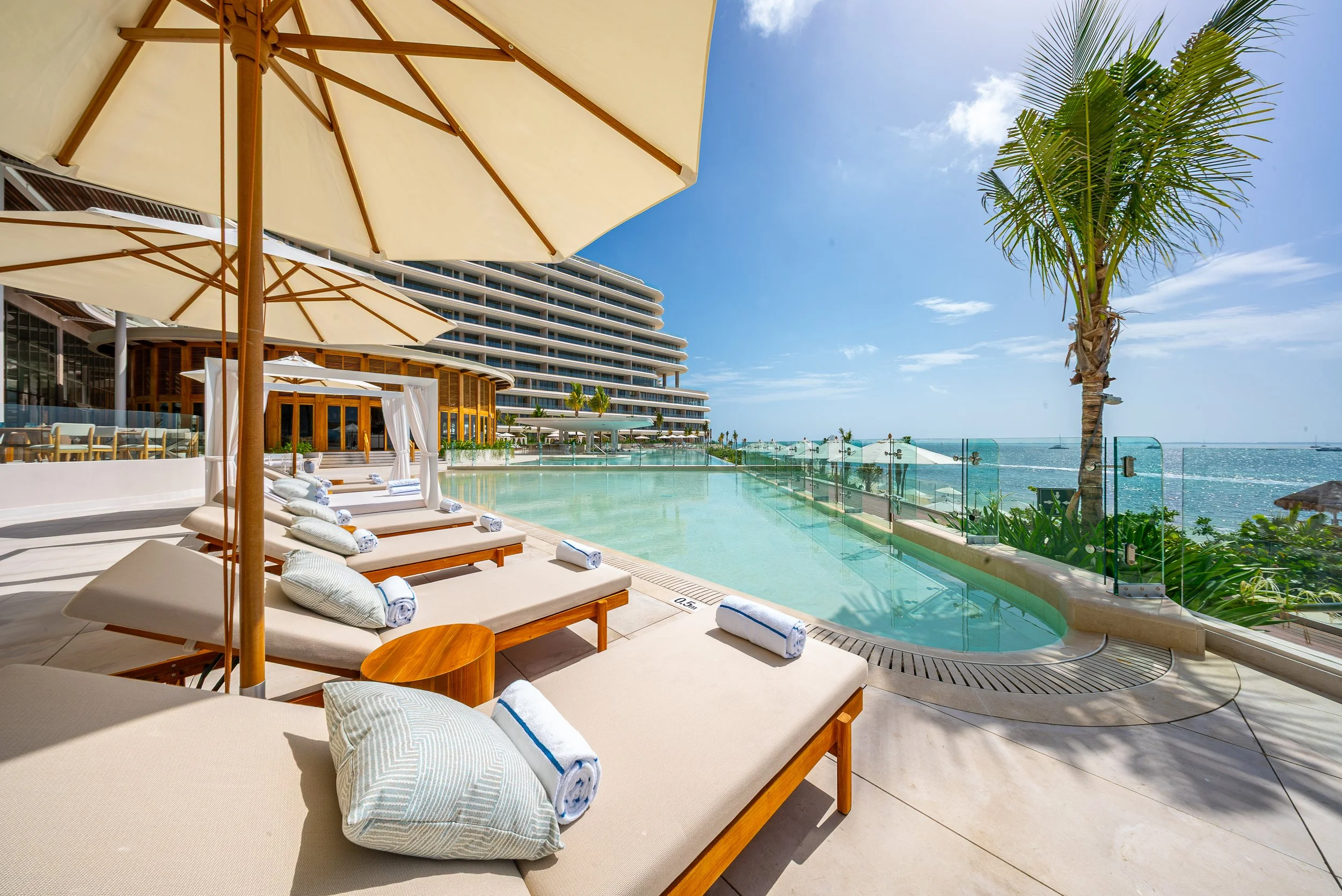 Poolside area with lounge chairs and umbrellas at a modern hotel with a building and palm trees overlooking the ocean on a sunny day.