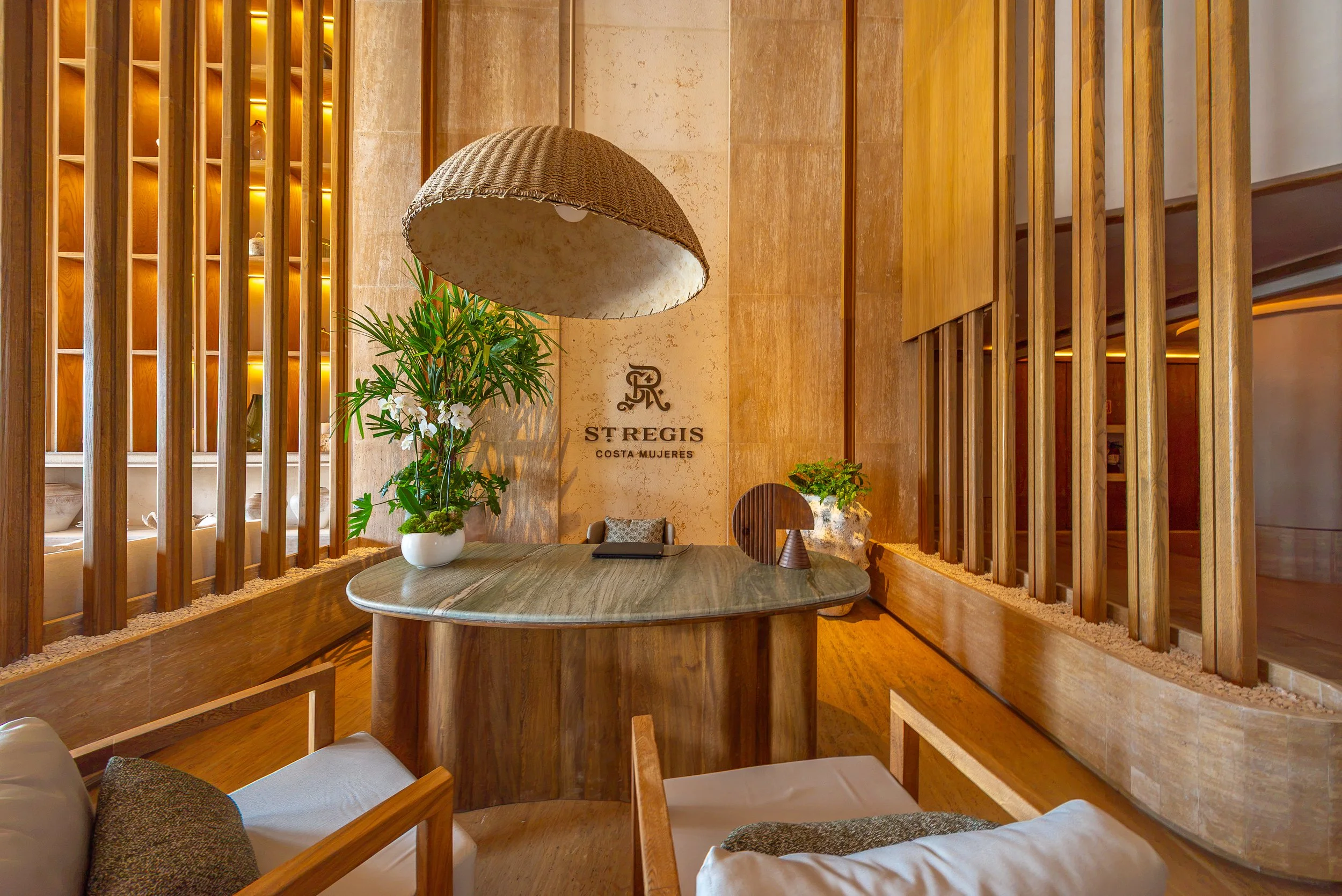 Interior of a hotel lobby with wooden room dividers, a wall lamp, plants, and seating area.