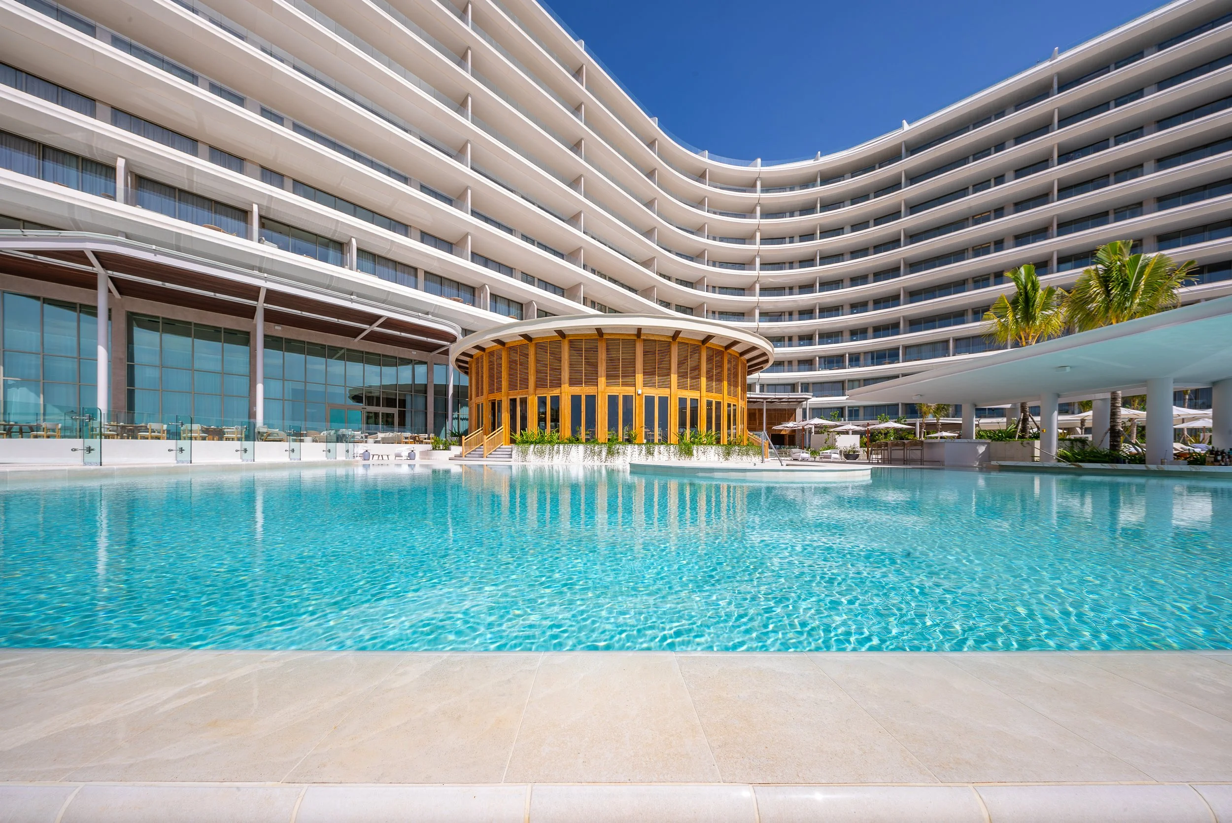 Luxury hotel with a large swimming pool in front, modern curved white building with glass balconies, a small wooden round building near the pool, and palm trees under a blue sky.