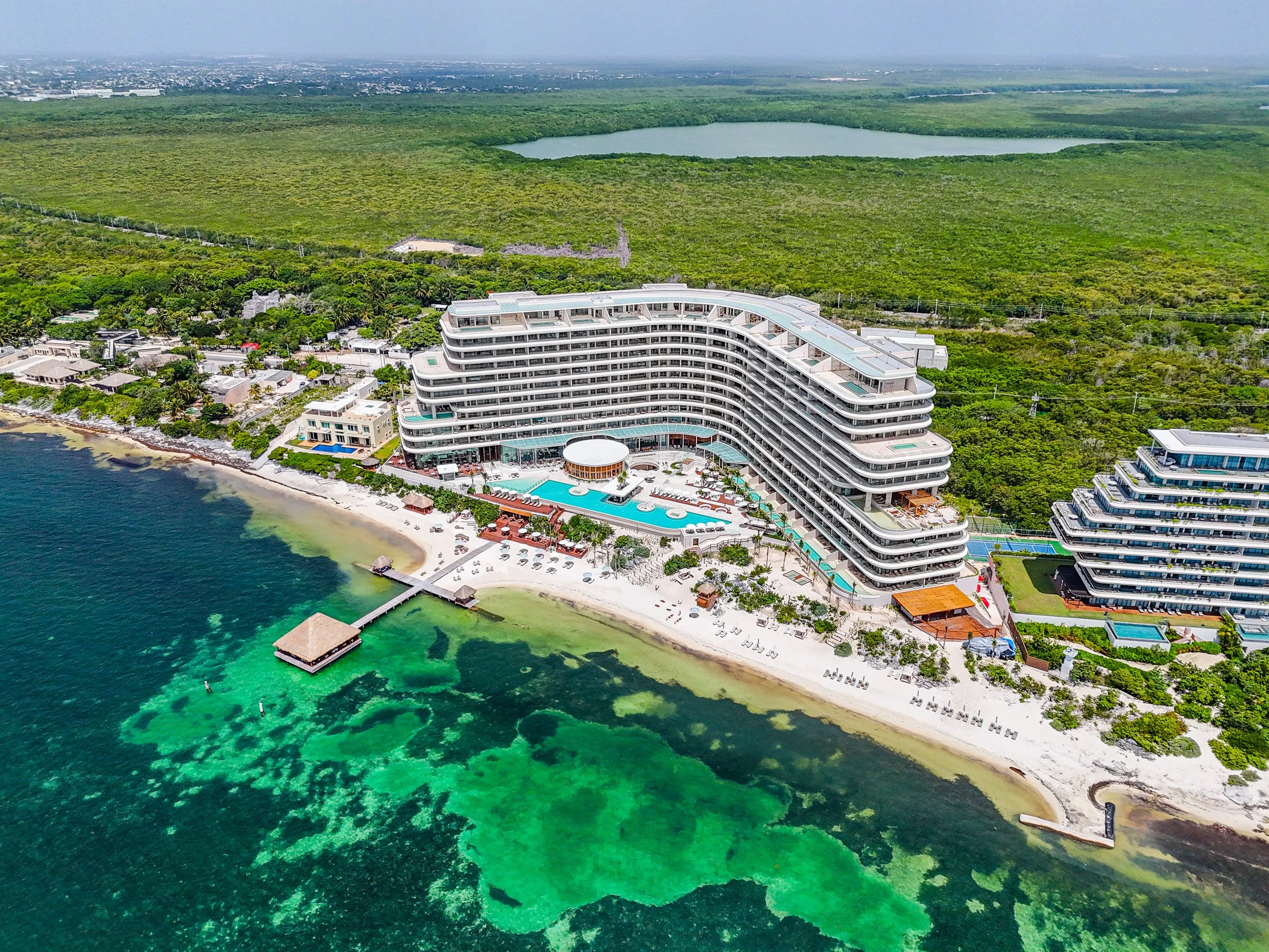 An aerial view of a large beachfront hotel with a pool, surrounded by trees and smaller buildings, with clear turquoise water and a sandy beach in the foreground, and a green forest and a lake in the background.