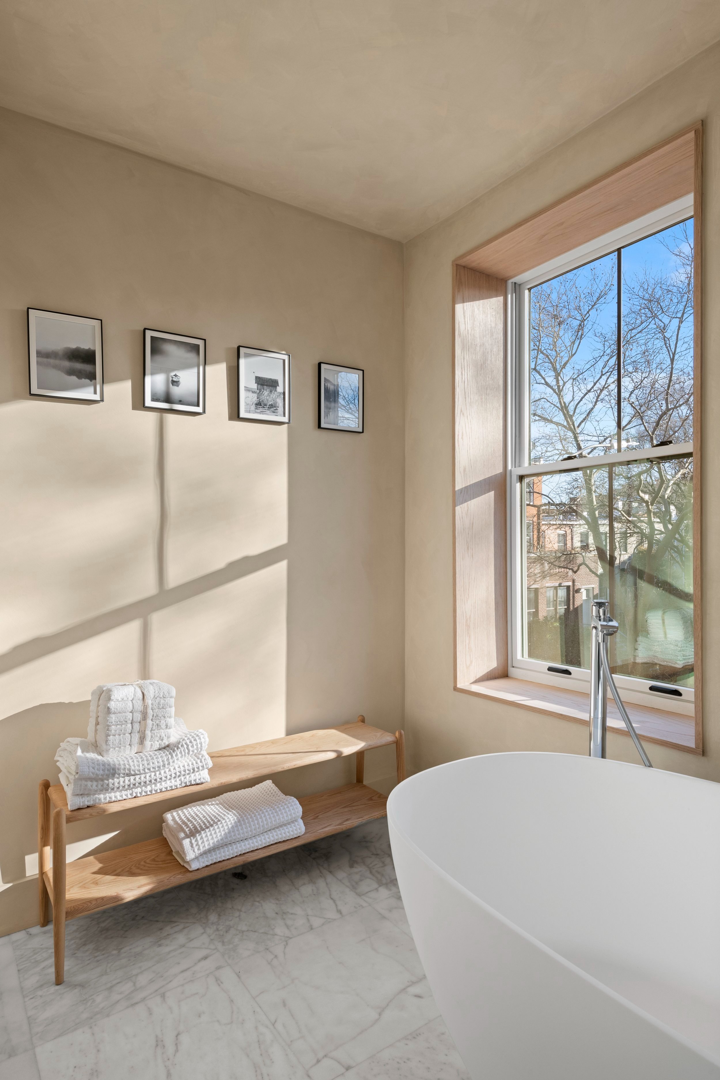 A minimalist bathroom with a freestanding bathtub, a wooden bench with folded towels, a large window letting in natural light, and black-and-white framed photos on the wall.