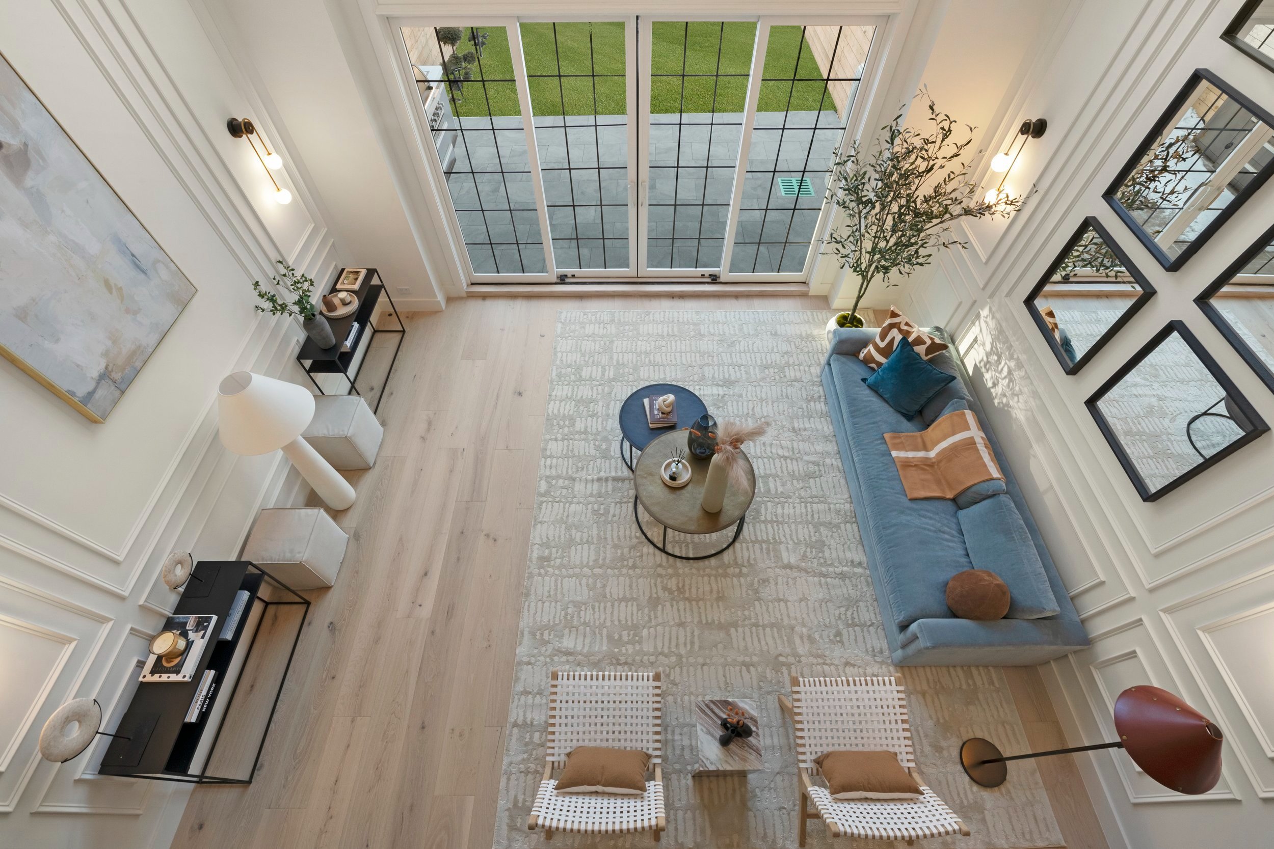 Living room with large sliding glass doors leading to an outdoor patio, hardwood floors, a blue sofa with decorative pillows, a beige area rug, two round coffee tables, a black console with decor items, a tall potted plant, three framed mirrors on th