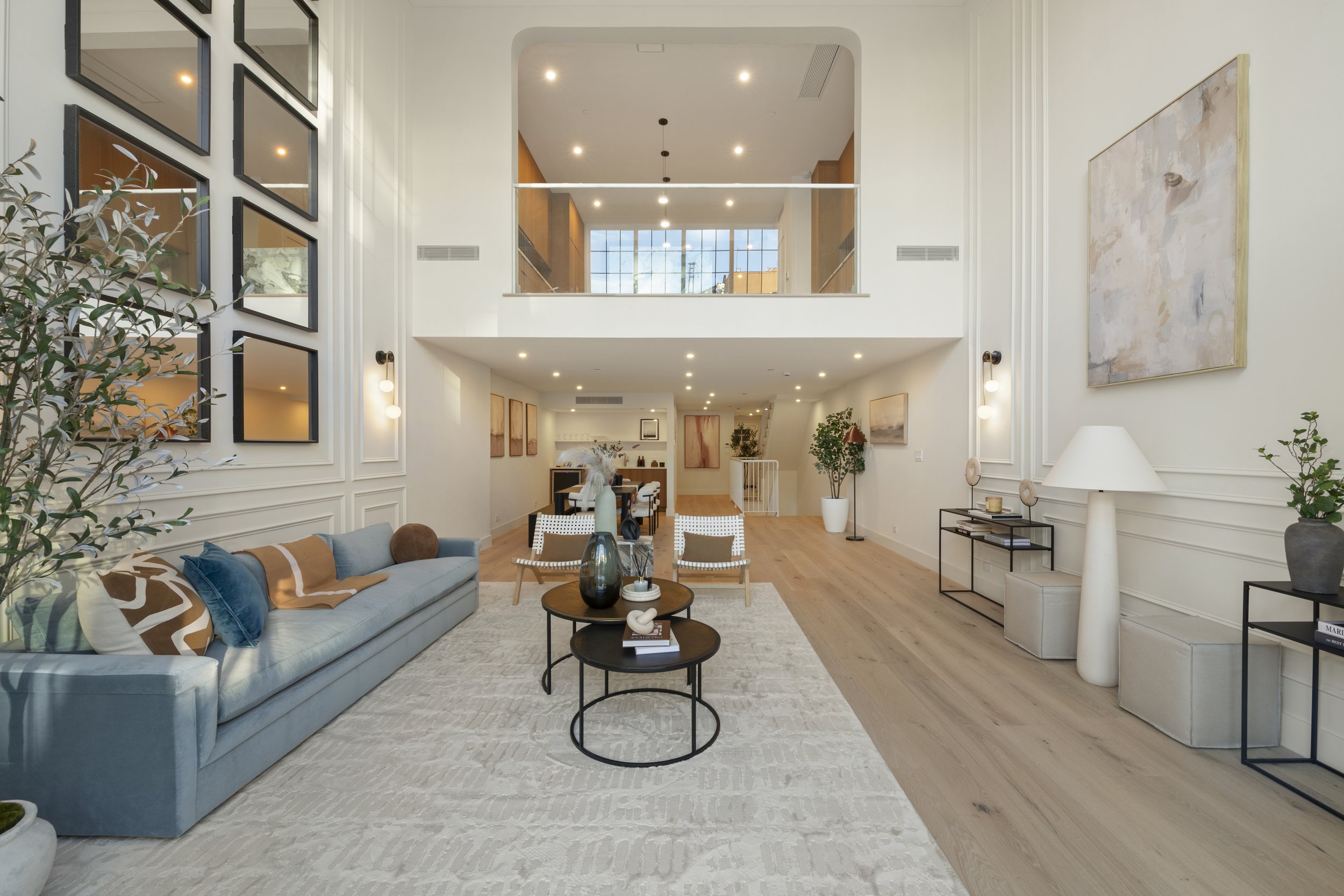 Spacious, modern living room with high ceilings, large windows, neutral-colored walls, light wood flooring, a long light blue sofa with pillows, and a pair of small round coffee tables with decorative items. Artwork and plants decorate the room.