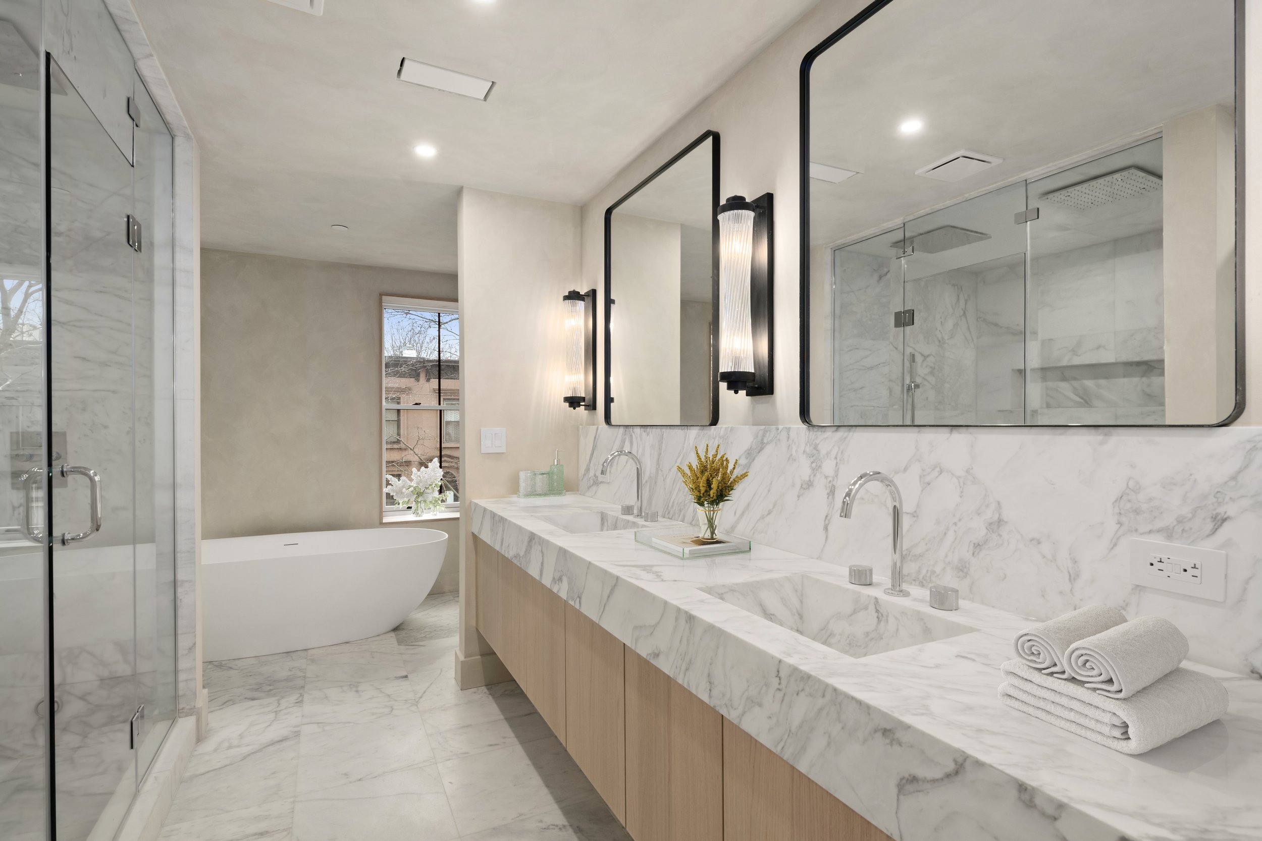 Modern bathroom with marble countertops, dual sinks, large mirrors, black light fixtures, a white bathtub, and a glass shower enclosure.